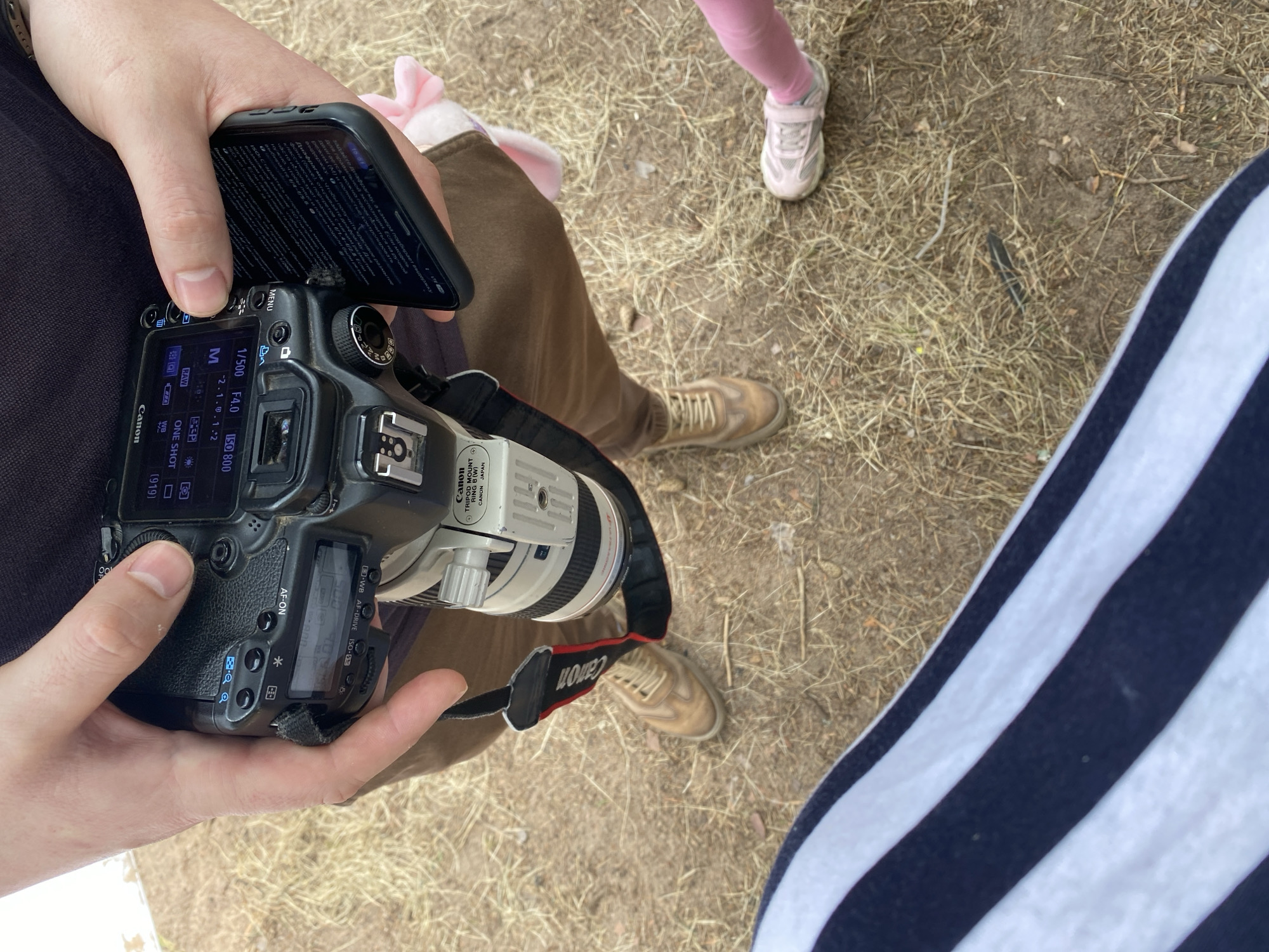 The image shows a close-up view of a person holding a Canon camera and a smartphone. The camera has a Canon brand lens attached. The person is wearing brown pants and tan shoes. A small child's pink leggings and sneakers are visible in the lower right corner of the image. The background appears to be an outdoor environment with dry grass and dirt. The image does not contain any information that would allow for its localization to Baltiiskoe, Russia.