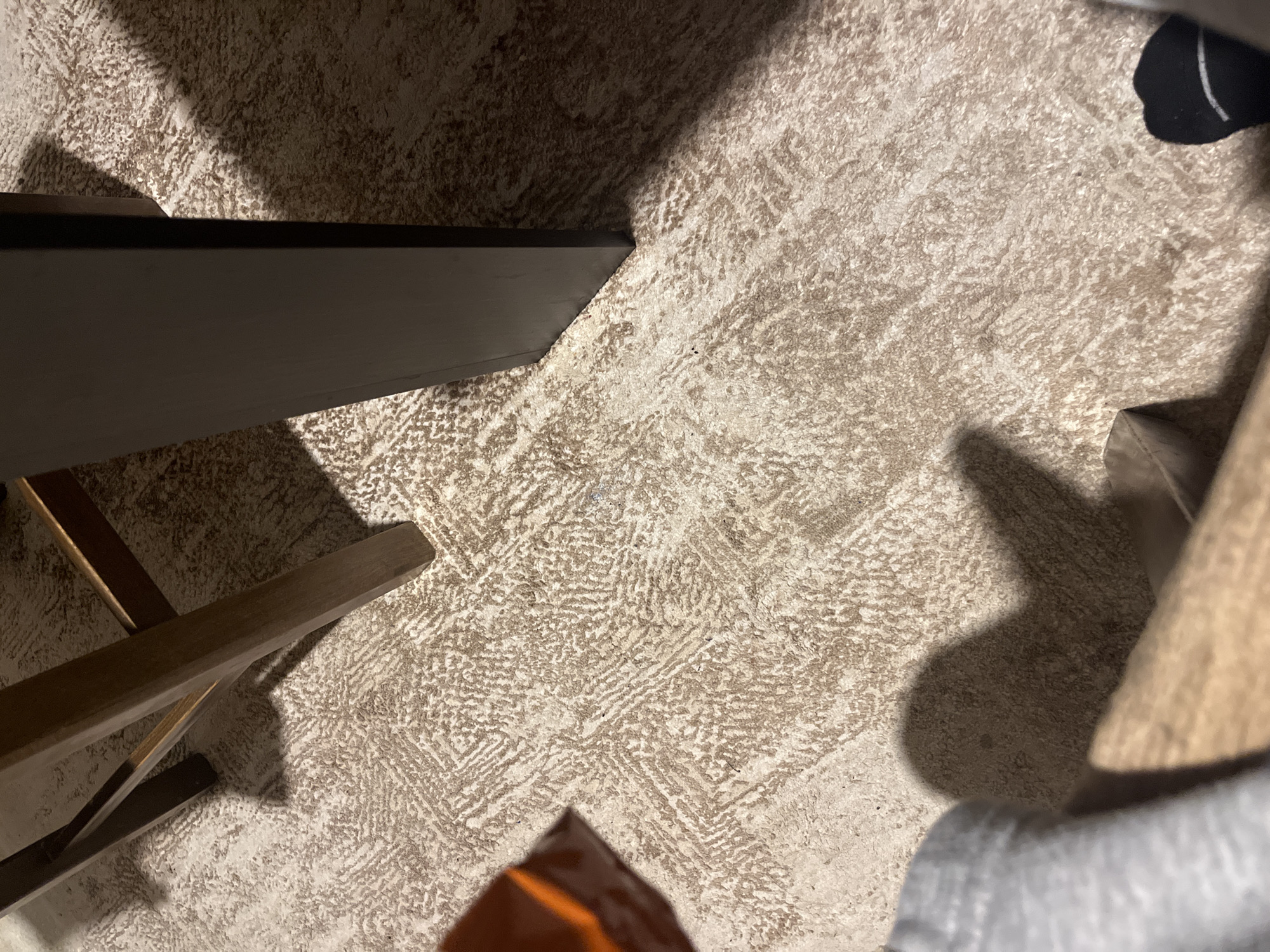 The scene, located in Blagoevgrad, Bulgaria, depicts an indoor environment focused on a light-colored, patterned carpet. The carpet features a subtle, textured pattern of irregular geometric or organic shapes in beige and light brown tones. In the upper left quadrant, a light brown wooden furniture leg, possibly from a chair or stool, is visible, along with darker wooden cross-members. A dark, flat, vertical furniture leg or panel, likely black or dark brown, is present along the right edge of the frame, extending vertically.
Strong, distinct shadows are cast across the carpet. A prominent, dark shadow from the dark vertical panel extends to its left. Another shadow, resembling the profile of a human foot and lower leg, is cast in the lower center of the image. In the lower right corner, a black sock with a white horizontal stripe is visible, indicating the presence of a person. A blurred, partially visible grey fabric, possibly clothing, is in the lower left. In the mid-left, a blurred object with an orange surface and a brown edge is partially visible. The perspective is downward-angled, capturing the floor and lower portions of objects and individuals.