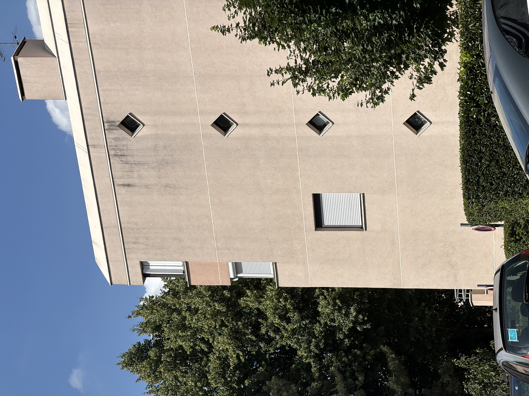 The image depicts the exterior of a building, possibly an apartment complex, on a clear, sunny day. The building's facade is a light, neutral color, with vertical lines dividing sections of the wall. A notable architectural feature is the presence of several diamond-shaped windows, arranged in a diagonal pattern. One larger rectangular window with blinds is also visible.
At the top of the building, a small, box-like structure with an antenna juts out. Lush green trees partially obscure parts of the building, with their branches reaching towards the facade. To the right of the building, a dense hedge runs along the side. The lower portion of a car is visible on the far right, suggesting a street or parking area. In the lower left of the frame, a large tree fills the space, contrasting with the clear blue sky.
The lighting suggests it is daytime, and the shadows cast by the trees and the car indicate a bright, sunny atmosphere. The scene appears to be in a residential or urban setting. No people are visible in the image. There is a small, circular sign with a red border and a diagonal line through it, often indicating a prohibition, visible on the hedge. A sticker with a blue background and white symbols is on the rear window of the car. The location context of Évry-Courcouronnes, France, places this scene in a specific geographical area.