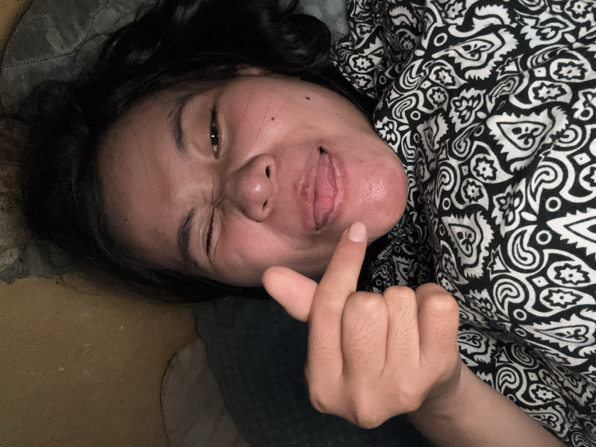 A close-up selfie shows a person lying down, with their head resting on a pillow. They are looking at the camera, winking one eye, and sticking out their tongue. Their right index finger is pointed towards their mouth. The person is wearing a black and white patterned shirt. The background appears to be a bedroom, with a yellowish wall visible behind them. The overall lighting suggests it is nighttime or a dimly lit room. There is no identifiable information to pinpoint the location within East Jakarta, Indonesia.