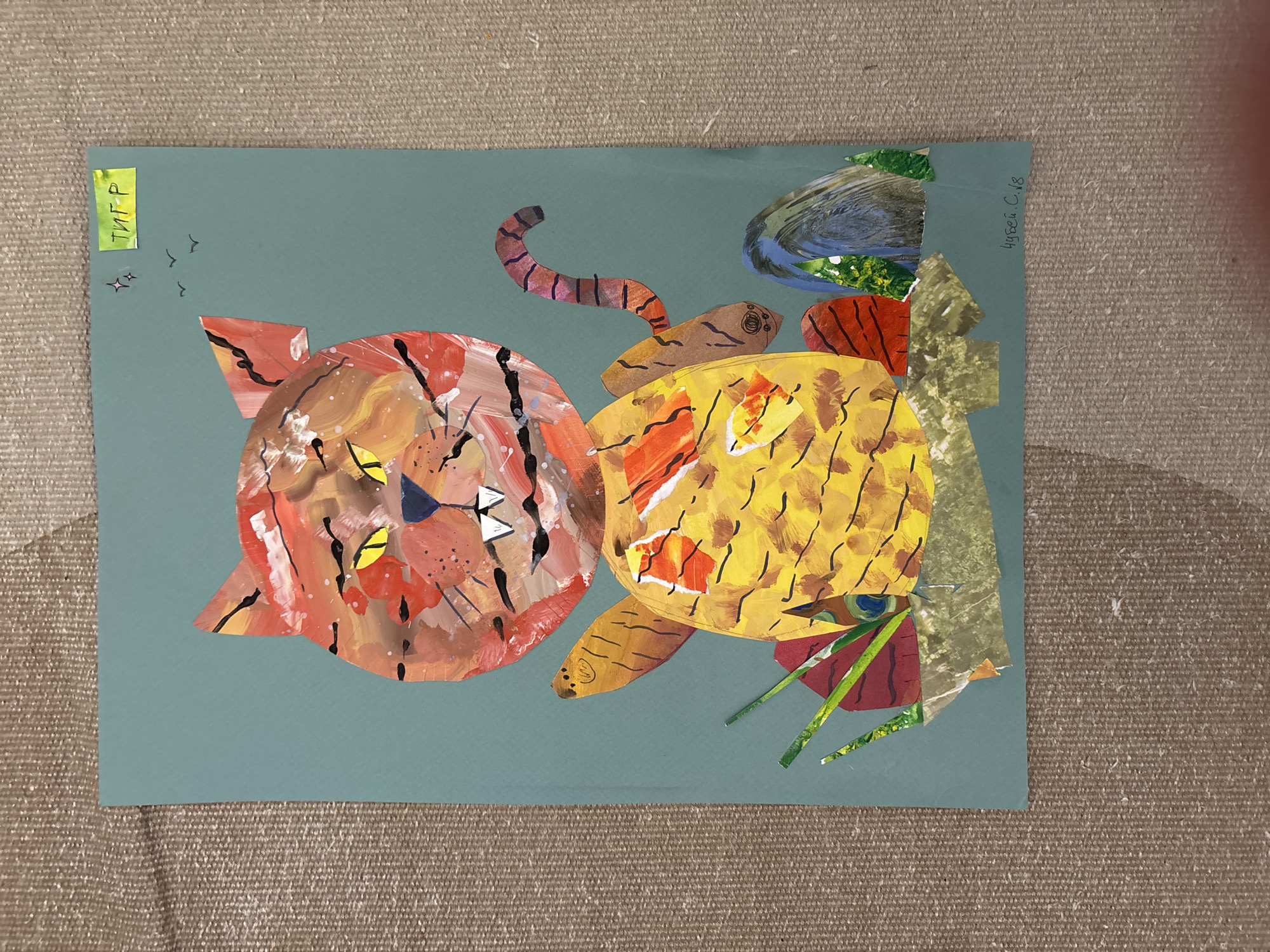 The image shows a child's art project featuring two cartoonish tigers. The tigers are constructed from painted paper circles and various scraps of paper. The left tiger is reddish-orange with black stripes and white triangles for whiskers, while the right tiger is yellow with orange and brown stripes. A segmented, curling tail extends from the right tiger. Additional paper scraps depict foliage, a bird, and other abstract shapes. The artwork is glued to a blue-green paper backdrop. "ТИГР" is written on a yellow label in the upper left corner, presumably the Russian word for "tiger". "Kygea C.18" is written in the upper right corner, likely the artist's name and year of creation. The artwork is resting on a textured beige surface, which is most likely the surface of a table. No information in the image implies a location in Saint Petersburg, Russia.