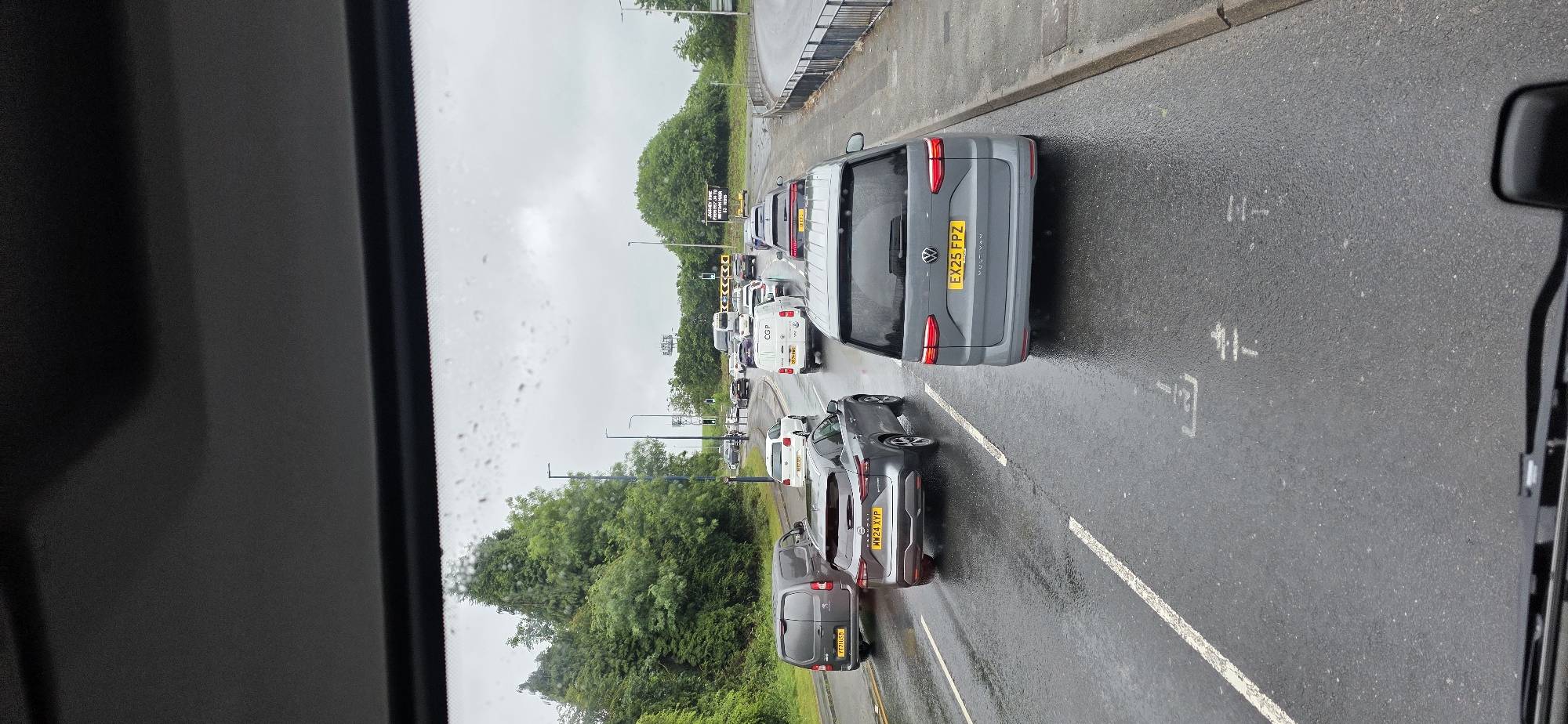 The image captures a traffic jam on a wet road in Tameside, United Kingdom. The scene is viewed from inside a vehicle, looking forward through the windshield, which is dotted with raindrops. Several cars and vans are visible on the multi-lane road, all stationary or moving very slowly.
In the foreground, a grey Volkswagen Multivan with license plate EX25 FPZ is prominent. To its left, a grey Nissan Qashqai with license plate MW24 XYP is also visible. Further to the left, a dark grey panel van with license plate TT21 HSB is in the adjacent lane. In the middle distance, more vehicles, including white vans and cars, are seen. A white van with "CGP" branding is also present. The road is wet, reflecting the overcast sky. Green trees and bushes line the left side of the road, while a metal fence and pavement are on the right. In the distance, traffic lights and signage are visible, indicating a junction or roundabout. The overall impression is of a typical congested commute on a rainy day in an urban or suburban setting.