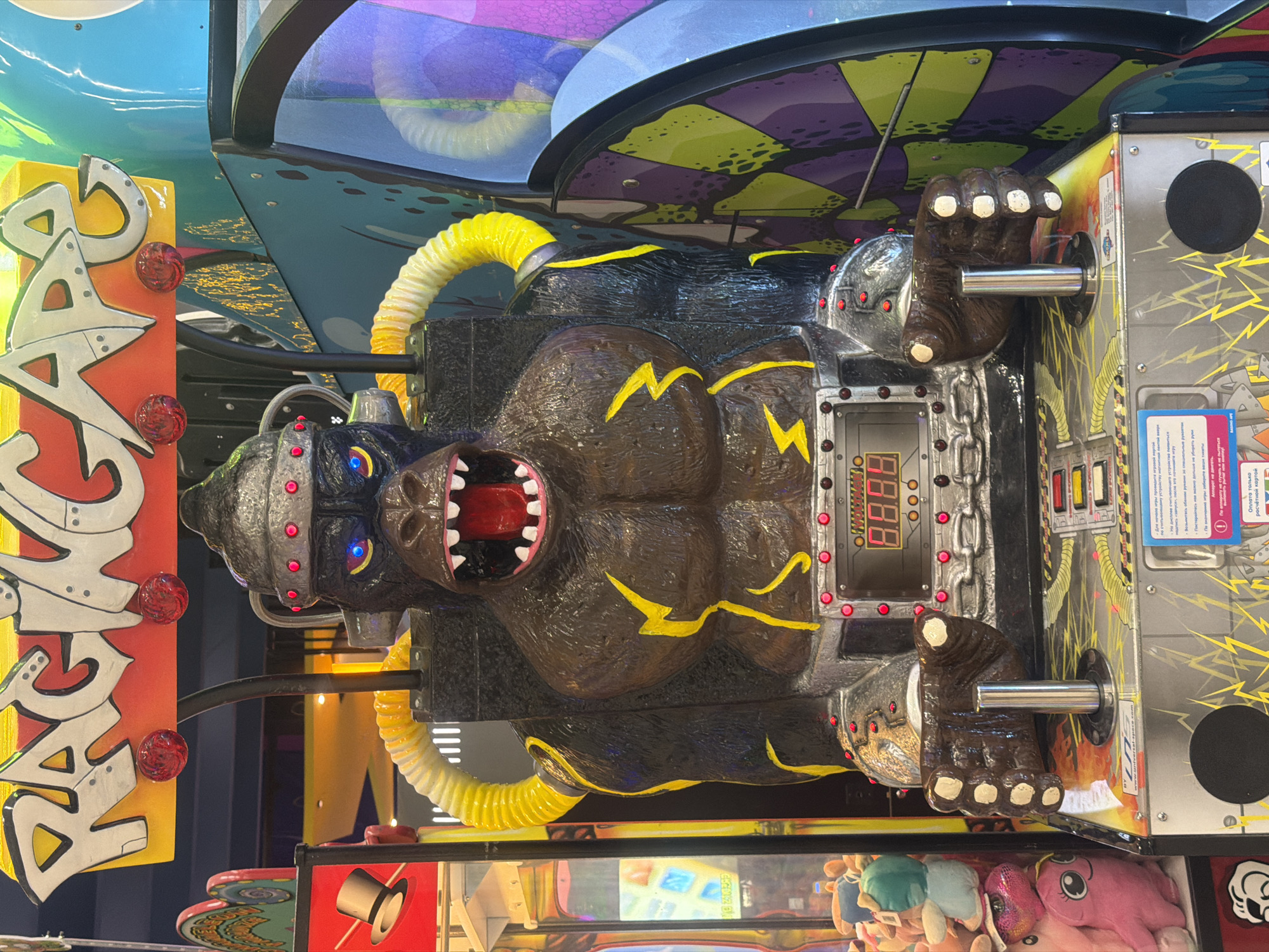 The image displays a "RAGINGAPE" arcade game featuring a prominent, dark brown, robotic gorilla figure. The gorilla wears a silver helmet adorned with red studs, has glowing blue eyes, and an open mouth revealing a red interior and white teeth. Yellow lightning bolt-shaped accents are present on its chest and arms, with yellow corrugated tubing extending from its shoulders.
A silver control panel on the gorilla's chest exhibits a red digital display showing "88:88" and surrounding red indicator lights. Below this, on the game's silver base, two dark brown gorilla fist-shaped controls, each with white knuckles, grip silver vertical joysticks. The base features yellow lightning bolt graphics and includes white labels with Russian text detailing game instructions and payment methods, indicating a Russian-speaking environment. A black circular vent is visible on the lower left of the base.
To the immediate left of the main game, a portion of a claw machine filled with various plush toys, including a pink octopus-like creature and other stuffed animals, is visible. To the right, another arcade machine with vibrant blue, purple, and lime green geometric patterns is partially observed. The scene is an artificially lit indoor environment, consistent with an arcade or family entertainment center, located in Perm, Russia. No individuals are present in the frame.