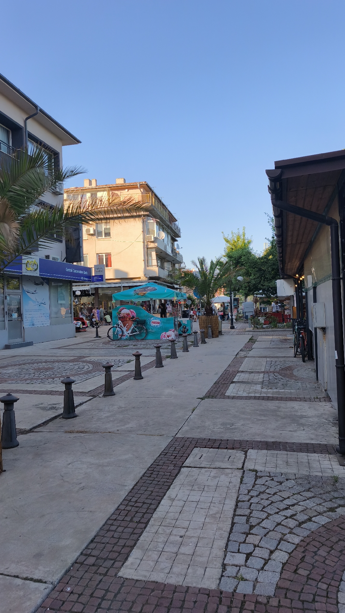 The image depicts a street scene in Pomorie, Bulgaria, likely during daytime with clear skies. The perspective is from a pedestrian walkway looking down a street lined with buildings on either side. The walkway is paved with a combination of large concrete slabs and smaller brickwork in a patterned arrangement. Numerous dark, bollard-like posts are spaced along the edge of the walkway.
On the left side of the street, a multi-story building with a modern facade and balconies is visible, partially obscured by a palm tree. Adjacent to it is a lower-rise structure with shops. One shop, identified as "Central Cooperative Bank," has a large glass storefront with informational displays. Further down this side of the street, an outdoor stall with a turquoise canopy and images of ice cream is set up, with people gathered around it. A bicycle is parked near the stall.
On the right side of the street, the edge of a building with a dark wooden overhang and string lights is visible. Further down the street, more buildings and trees can be seen. Another bicycle is parked near the building on the right. The street appears to be a commercial or pedestrian area, with several individuals walking or standing, suggesting a relatively public and active space. The overall ambiance suggests a warm, sunny day in a coastal town.