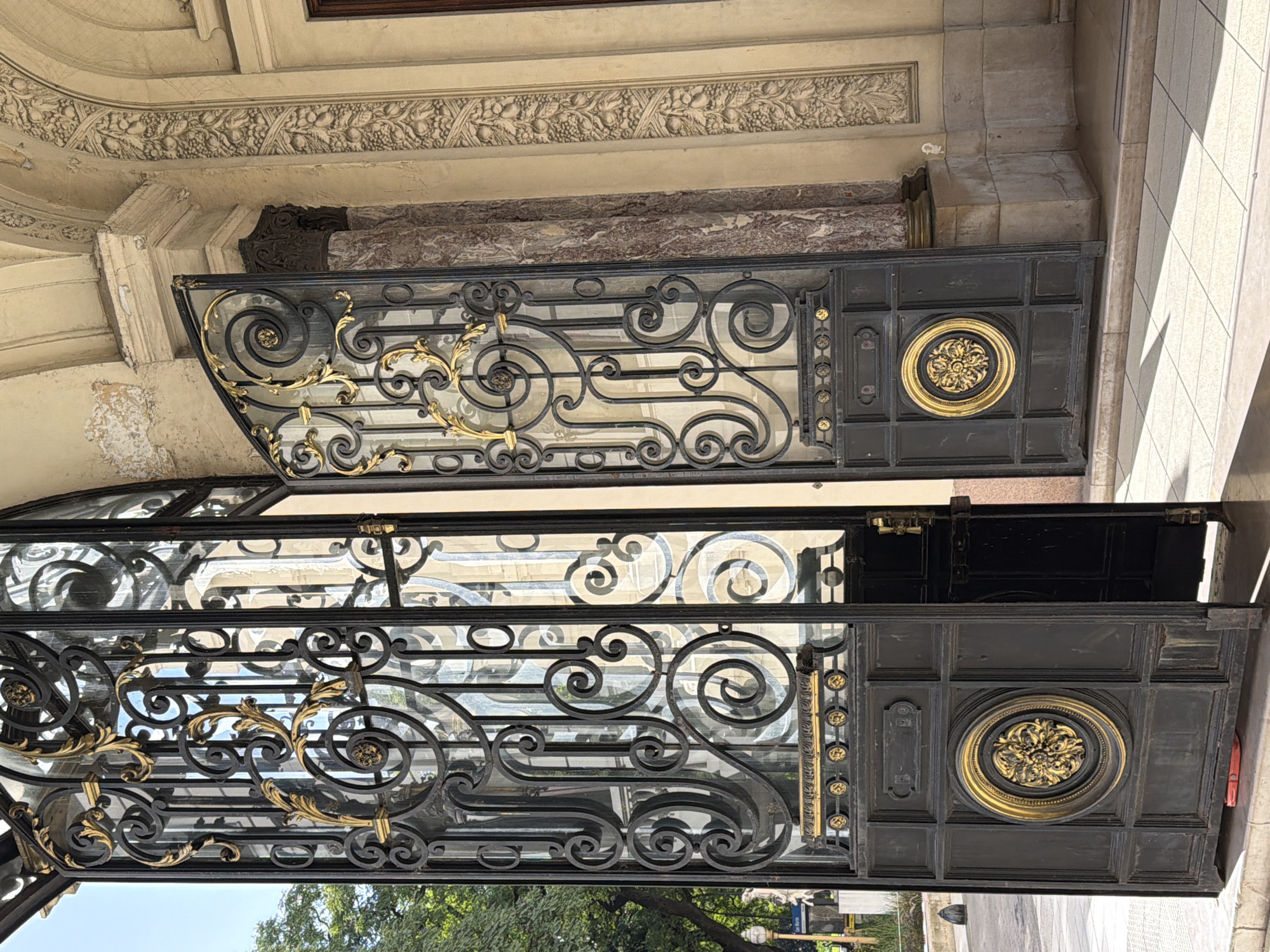 A pair of ornate, dark metal gates, embellished with gold-colored floral designs and circular medallions, are partially open, revealing glimpses of a tree-lined street scene beyond. The gates are attached to a building featuring detailed architectural elements such as decorative molding and marble accents. The scene appears to be located in Buenos Aires, Argentina, based on architectural style.