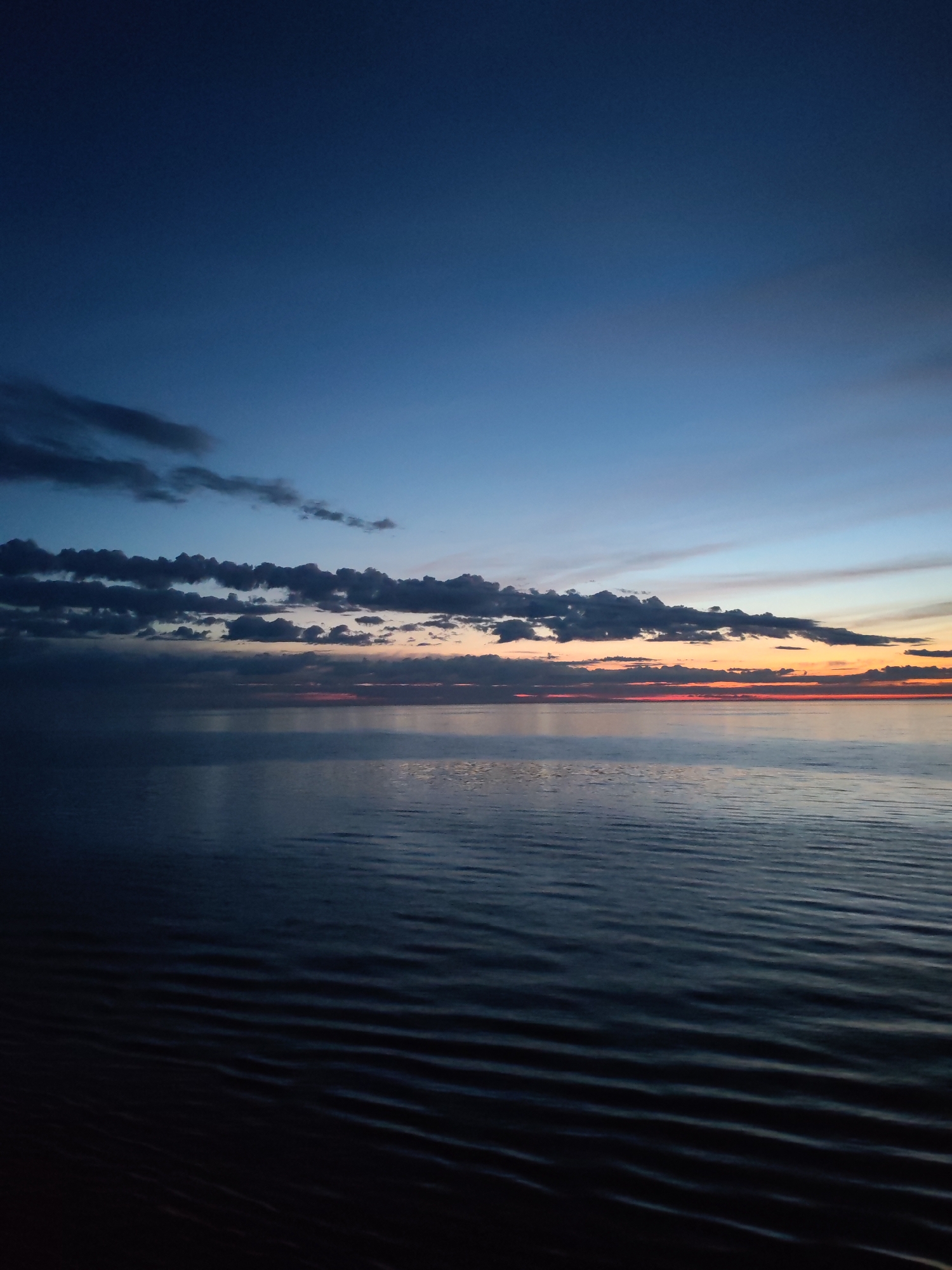 The image depicts a serene body of water, possibly a sea or ocean, under a dusky sky at twilight. Dark blue dominates the upper portion of the frame, transitioning into lighter blues near the horizon. A thin band of orange and red hues stretches across the horizon line, indicating the setting sun. A line of cumulus clouds is visible above the sunset line. The water's surface exhibits gentle ripples, creating a series of dark and light lines extending from the foreground towards the horizon. The scene appears devoid of any human presence or artificial structures. The location and city are unknown.