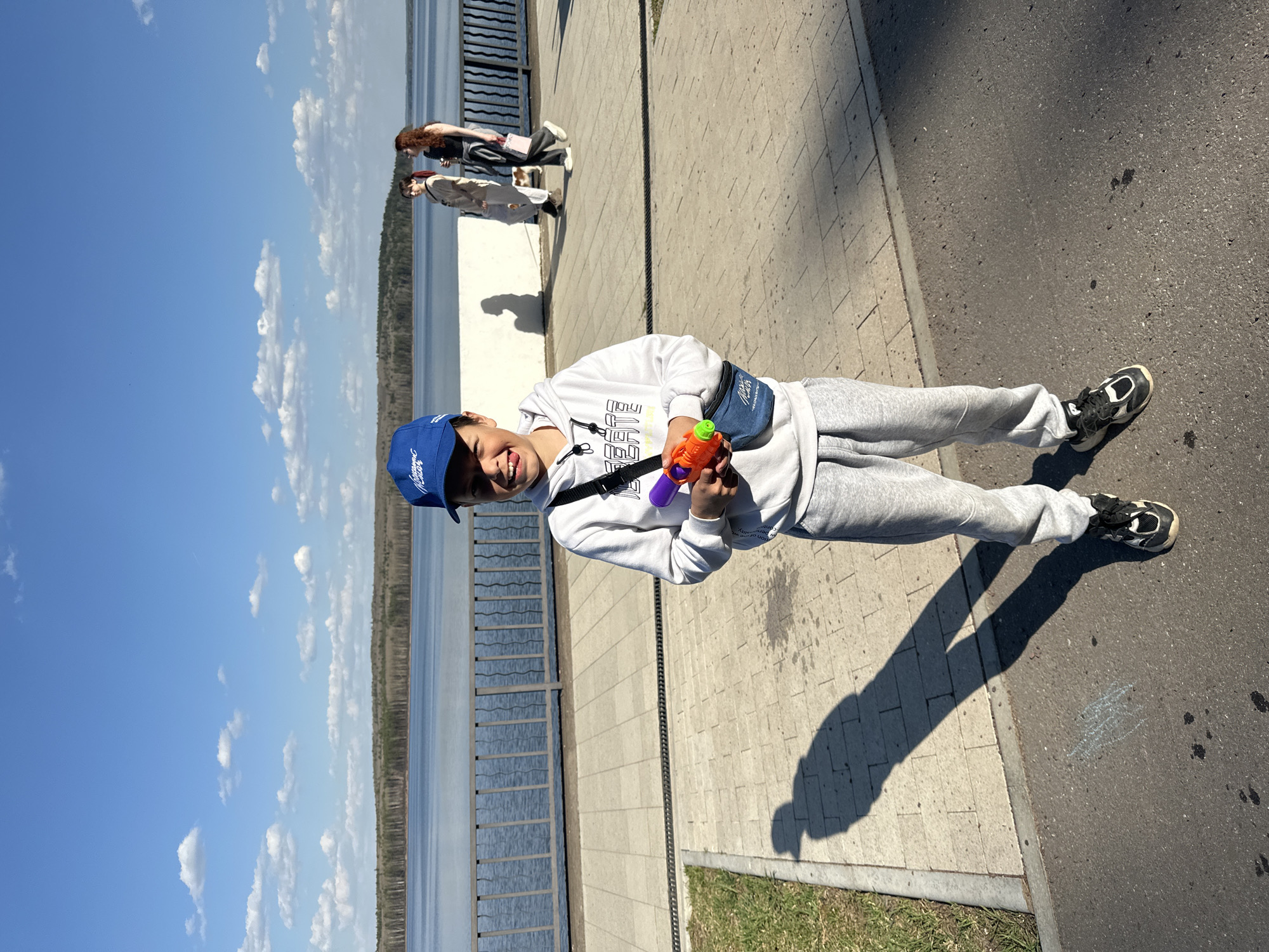A young person is standing on a sidewalk next to a railing, holding a colorful water gun. The person is wearing a light-colored sweatshirt, grey sweatpants, sneakers, a blue baseball cap, and a small bag. The person is smiling at the camera. In the background, there is a body of water, possibly a reservoir, and trees. Further back, there are two people standing near the railing. The scene appears to be located in Chaikovsky, Russia. The environment is sunny. The sidewalk is composed of rectangular paving stones and there is a strip of grass beside it. Next to the sidewalk, there is a road.