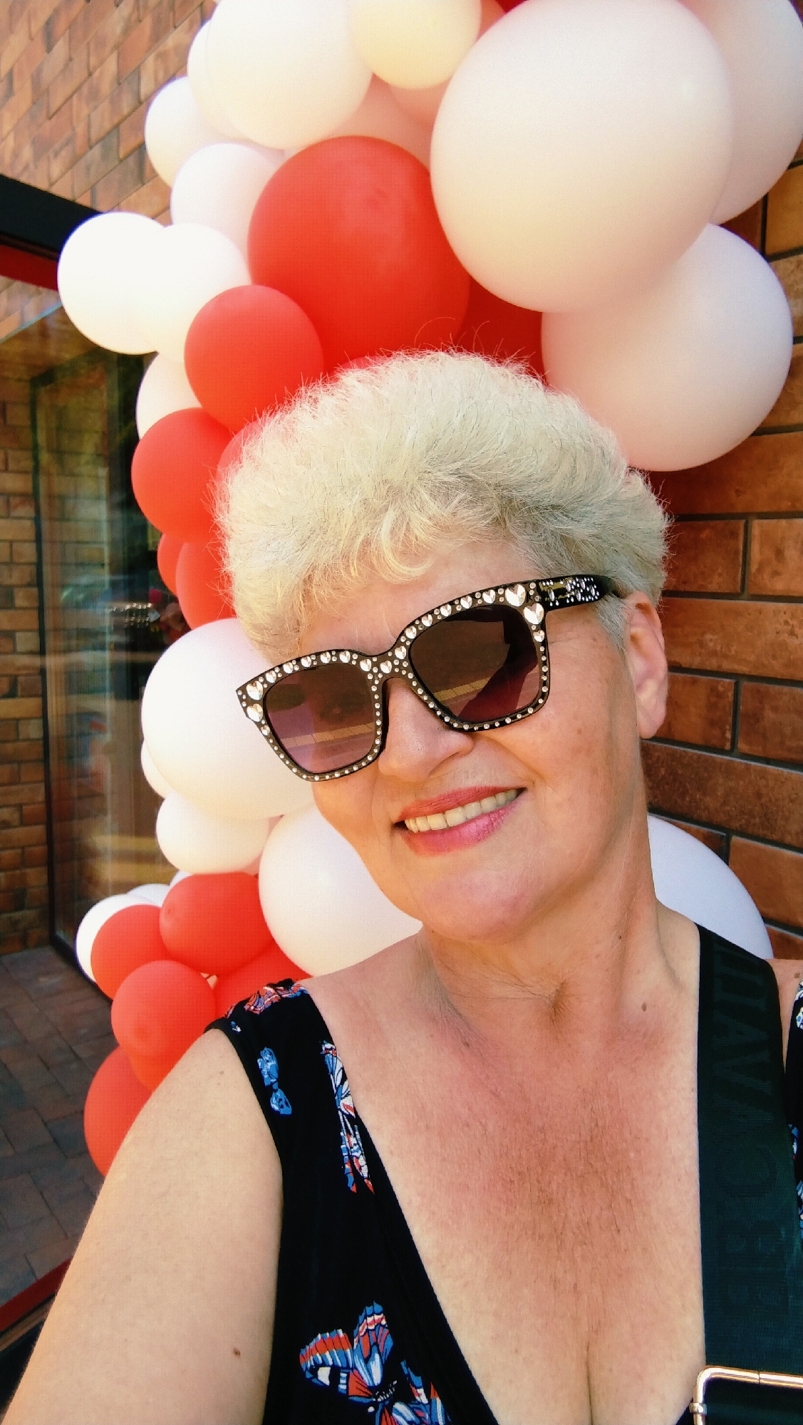 A medium shot photograph depicts a woman with short, permed blonde hair, smiling and wearing large, ornate sunglasses with a black frame embellished with rhinestones and hearts. She is positioned in front of a backdrop of red and white balloons, with a brick wall visible to her right and a glass storefront to her left, suggesting an outdoor or semi-outdoor location. The woman is wearing a black dress with a butterfly print. The scene is likely a celebration or event. The image is undated and its specific location within Horishni Plavni, Ukraine, is not provided.