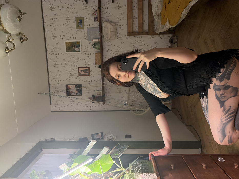 A woman with tattoos, including one of a woman's face on her thigh, poses in front of a mirror taking a selfie. She wears a dark outfit and holds her phone up. The background shows a room with patterned wallpaper, framed pictures, a hanging macrame chair, and a plant with grow lights. The floor appears to be hardwood. A vintage-style chandelier is partially visible at the top left. The overall image is a bit rotated and slightly blurry.