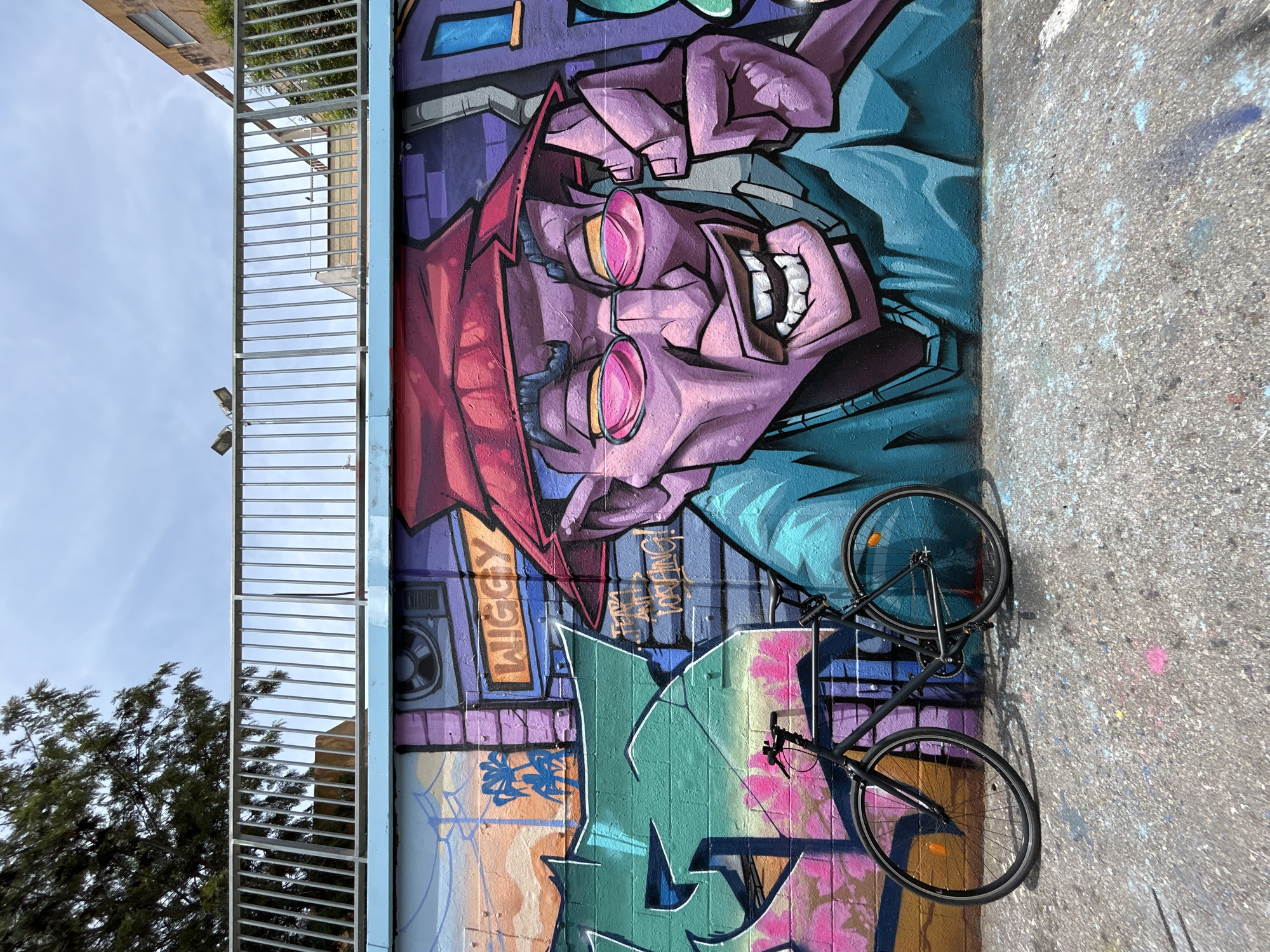 The image shows a close-up view of a colorful mural on a textured concrete wall. The mural features a stylized, cartoonish face with purple skin, large pink-rimmed glasses, and an open mouth revealing white teeth. The face is wearing a red head covering and a blue garment. Below the face, the mural includes abstract shapes in green, pink, orange, and blue, with some graffiti-style lettering. To the left of the mural, a metal railing is visible, with some trees and a building in the background. Leaning against the mural is a black bicycle, with its wheels clearly visible. The scene appears to be outdoors during the daytime, with bright sunlight casting shadows. The overall mood of the artwork is energetic and vibrant. Visible text on the mural includes "WIGGY" in an orange rectangle and some scribbled writing that is difficult to decipher. The location is identified as Barcelona, Spain.