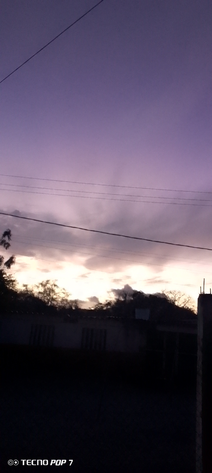 The image shows a landscape taken at dusk or dawn in Altagracia, Venezuela. The sky is a mix of purple and cloudy, with several power lines crossing the frame. In the middle ground, there are silhouettes of trees and a building with visible windows. The foreground is mostly dark, obscuring details, with the edge of a structure visible on the right side. The 'Tecno Pop 7' watermark is in the bottom left corner.