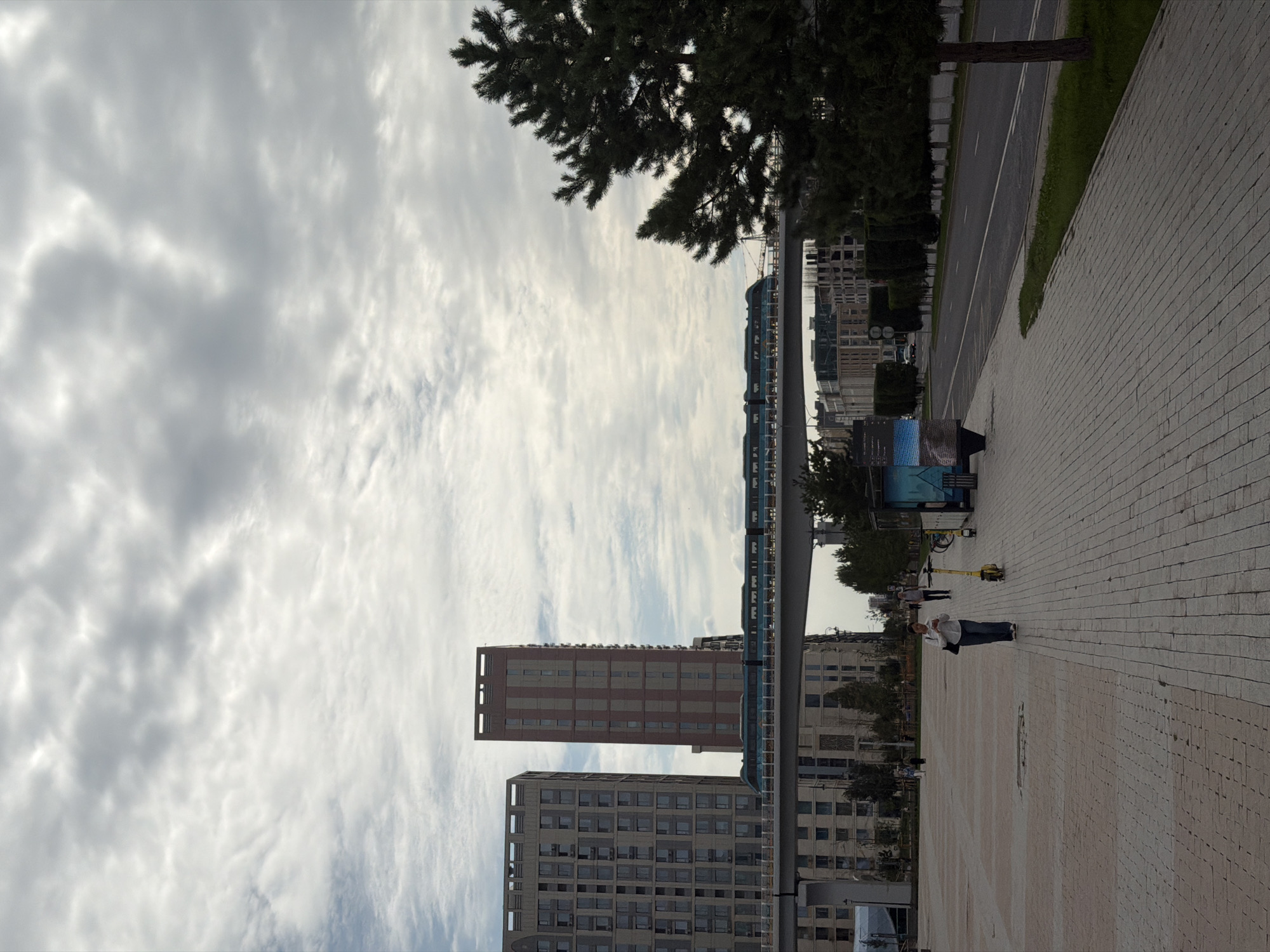 This is a landscape photograph of a modern urban environment in Astana, Kazakhstan, under a cloudy sky. In the foreground, a wide paved walkway, constructed of rectangular bricks, stretches into the distance. A person in a white shirt and blue jeans walks on the walkway, looking at their phone, with a dark bag slung over their shoulder. To their left, a yellow electric scooter is parked. In the mid-ground, several modern residential buildings with geometric facades are visible. A monorail train with a teal and dark blue exterior travels on an elevated track that spans between two buildings. The background features more urban architecture, including buildings with similar modern designs, and a row of manicured trees lining a street. A dense evergreen tree is positioned on the right side of the frame, partially obscuring the view of the background. A digital advertising screen is visible on the left side of the walkway. The overall atmosphere is overcast and cool.
