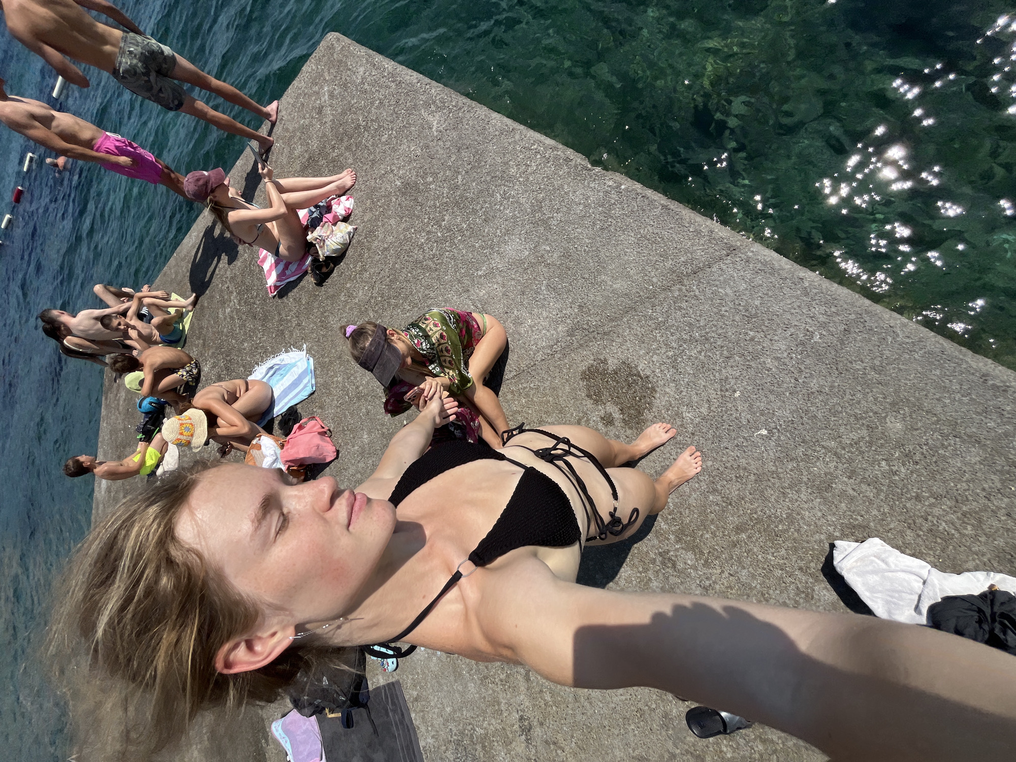The image shows a group of people sunbathing on a concrete surface next to clear, turquoise water. The scene is outdoors, suggesting a warm, sunny day, likely in the late morning or early afternoon given the bright sunlight casting distinct shadows. The water is calm and reflects the sunlight, creating a shimmering effect.
Several individuals are visible. In the foreground, a person is taking a selfie, looking towards the camera with their eyes closed, appearing relaxed. They are wearing a black bikini. Behind them, other people are scattered across the concrete. Some are lying down, while others are sitting or crouching. One person is wearing pink shorts and a hat, and another is in a patterned top and green bikini bottoms. A few are partially obscured or seen from behind.
Objects present include towels, bags, and what appear to be swimsuits. The concrete surface seems to be a pier or a waterfront area, with the edge of the water clearly visible. The overall atmosphere suggests a leisurely day spent by the water, possibly at a beach or a lake. There are no visible text elements in the image.