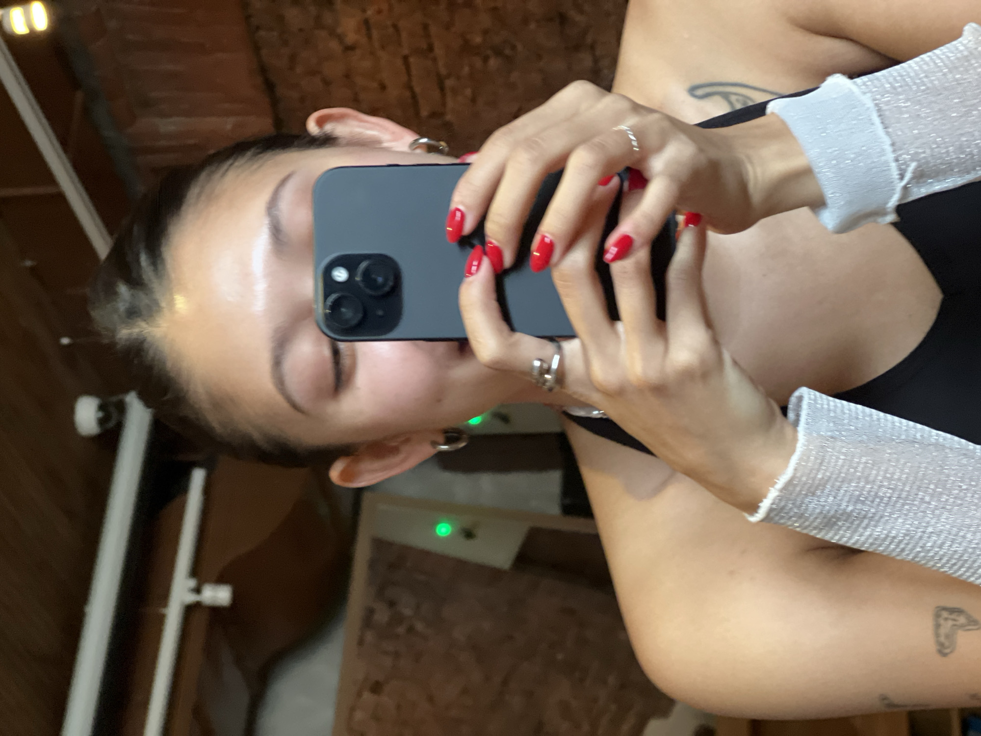 A woman is taking a selfie with a grey smartphone. She has fair skin, dark hair pulled back, and red nail polish. She is wearing a black top with sleeves made of white, sparkly material. The location appears to be indoors with a brick wall visible in the background. There is a metal rod and a shelf-like structure also visible in the background. There is no evidence indicating that the location is in Vladivostok, Russia.