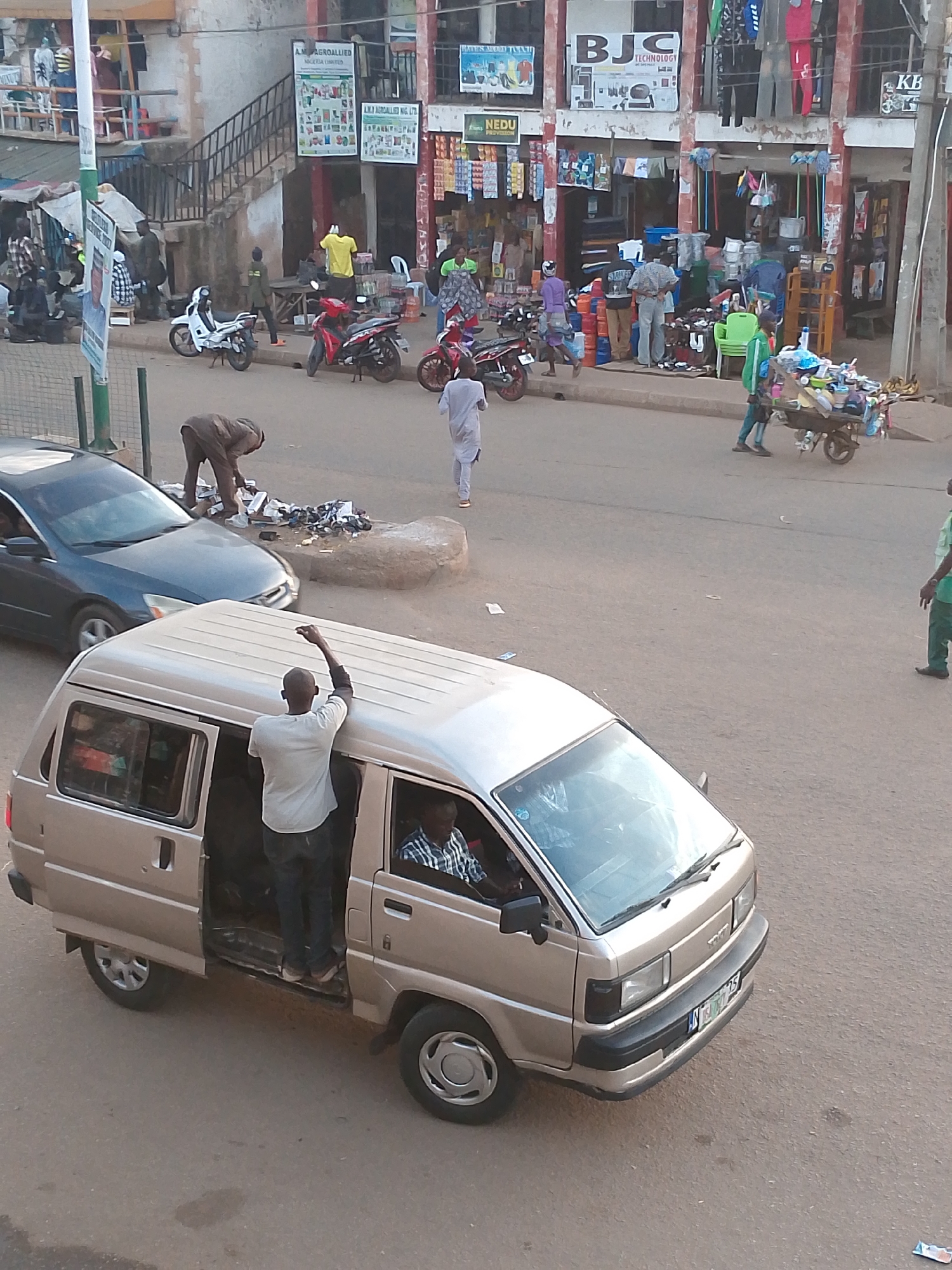 This is an outdoor, high-angle, eye-level shot of a bustling street scene in Bukuru, Nigeria. The foreground features a beige van with its sliding door open, revealing two individuals inside. One man, wearing a light-colored shirt and dark pants, stands in the open doorway with his arm raised. Another man, wearing a plaid shirt, is seated in the driver's seat. A dark blue sedan is parked to the left of the van, and a man in a brown outfit is bent over near the sedan, apparently tending to items on the ground.
In the background, various shops line the street, displaying merchandise such as clothing, cleaning supplies, and household goods. People are seen walking and interacting, with several motorcycles parked along the roadside. A man pushes a cart laden with various items across the street, and other individuals are visible near the shops. The overall impression is one of a vibrant commercial area with ongoing daily activity.