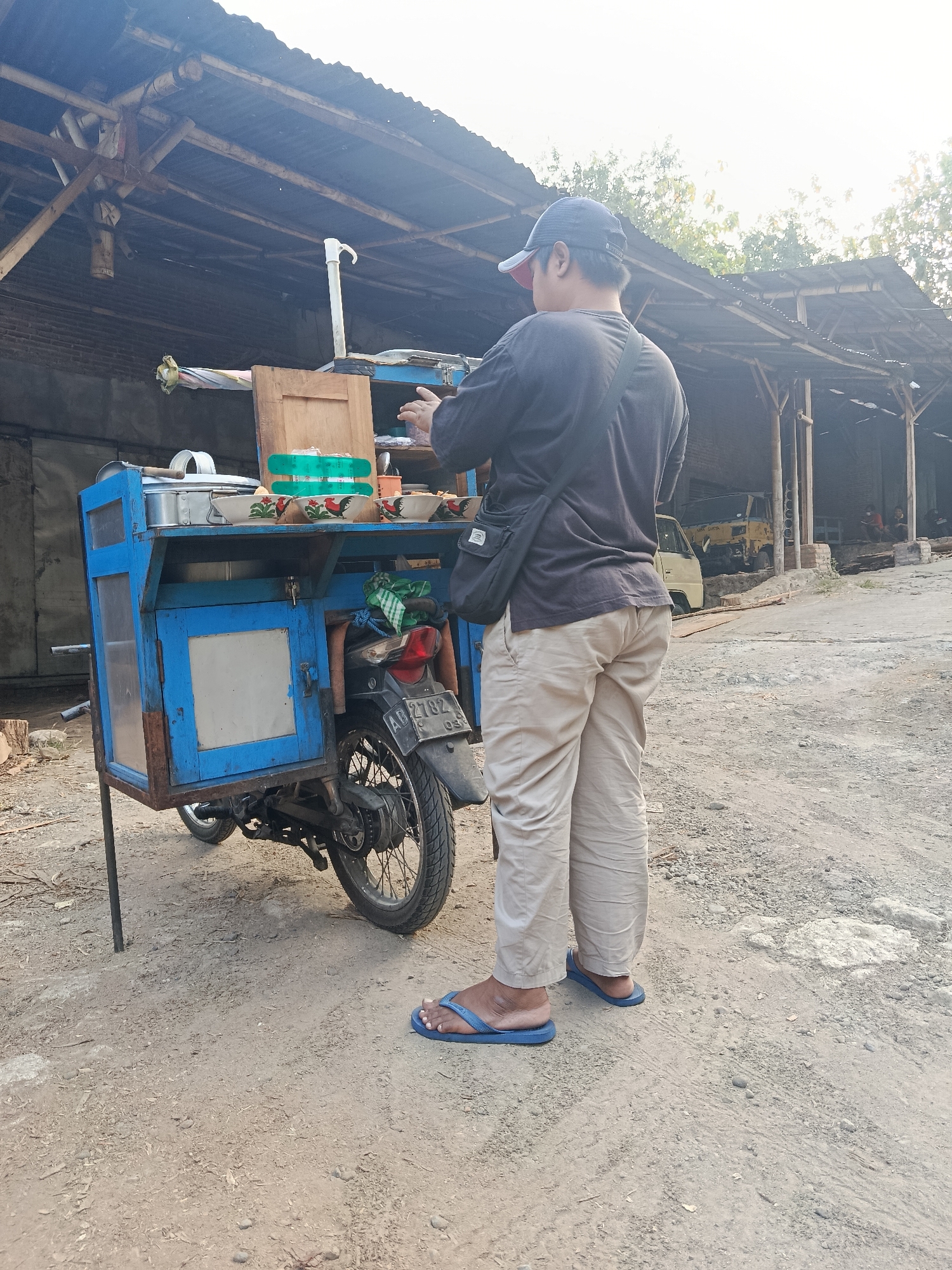 A person wearing a dark t-shirt, beige pants, a dark baseball cap, and blue flip-flops stands next to a blue and wooden food cart attached to a motorcycle. The food cart contains several bowls, a metal container, and various other items. The person appears to be interacting with the cart. The scene is outdoors, with a rustic, covered structure in the background and a dirt and gravel ground. The lighting suggests it is daytime, possibly late afternoon. The license plate on the motorcycle reads "AB 2702". The location is identified as Kasihan, Indonesia.
