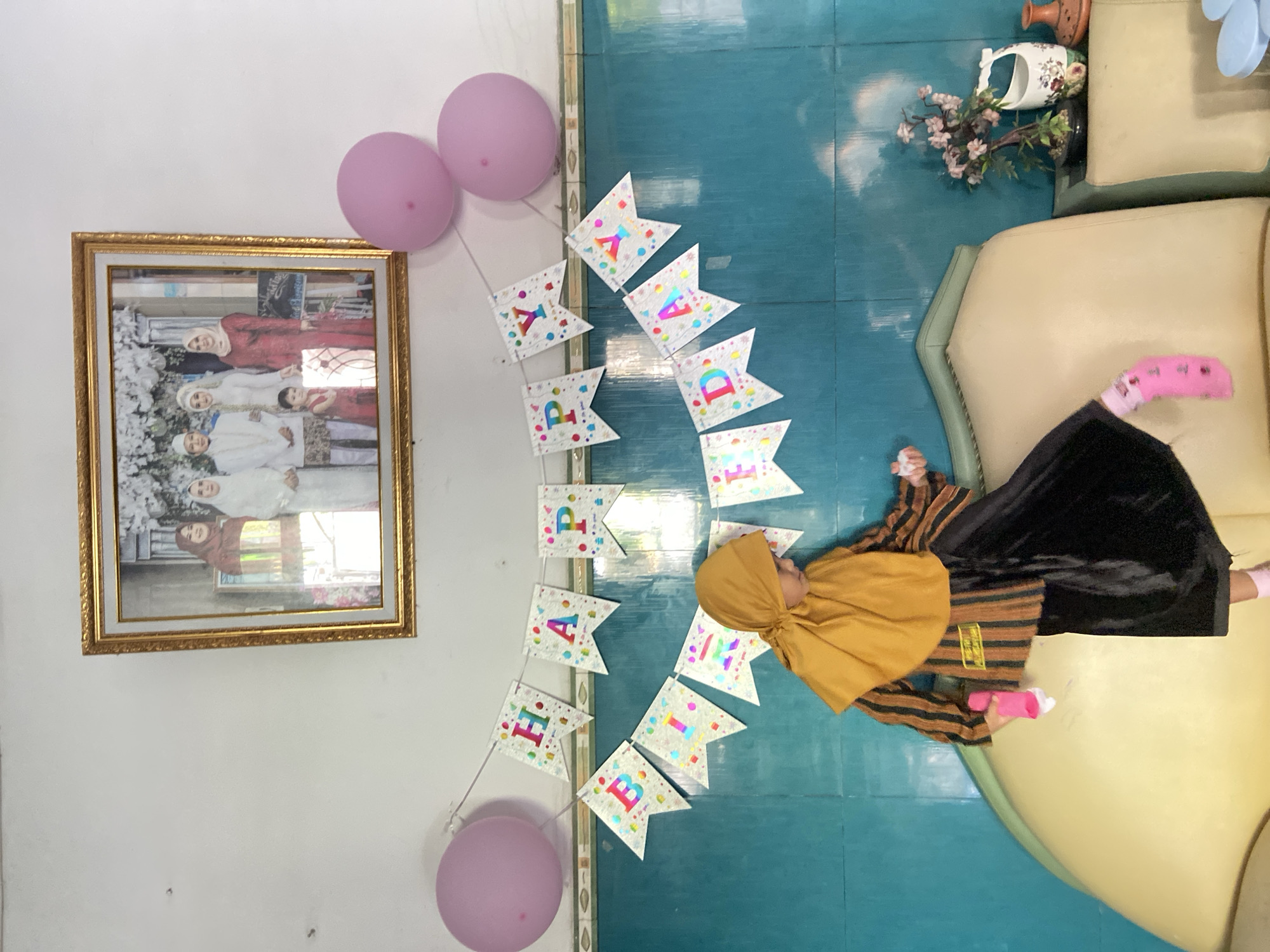 A child in a hijab and dress lies on a beige upholstered sofa, with pink socks on their feet. They are holding a pink object. Above them, a "Happy Birthday" banner is strung between two pink balloons. Adjacent to the banner is a framed photograph of a family group on a white wall. The floor is blue. There are decorative items near the sofa. This is Gelam, Indonesia.