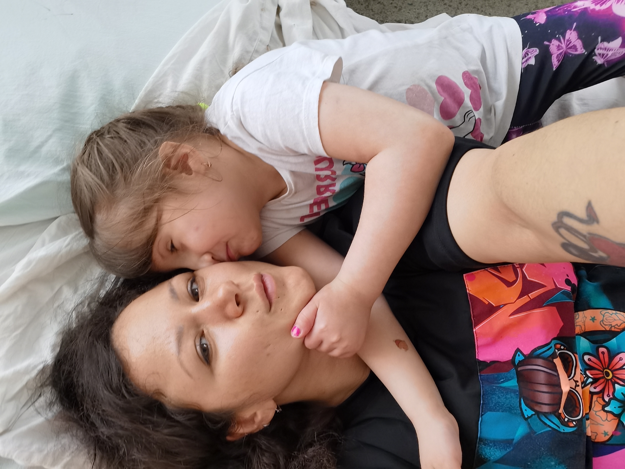 A young child and an adult female are lying on a light-colored fabric surface, likely a bed or blanket. The child, wearing a white t-shirt with pink designs and butterfly-printed leggings, is positioned partially on top of the woman, with the child's arm around the woman's neck and face close to the woman's cheek. The woman, with dark hair, is wearing a black top. Part of the woman's tattooed arm is visible. The woman is looking at the camera. The image does not provide visual information related to the city of Perm, Russia.