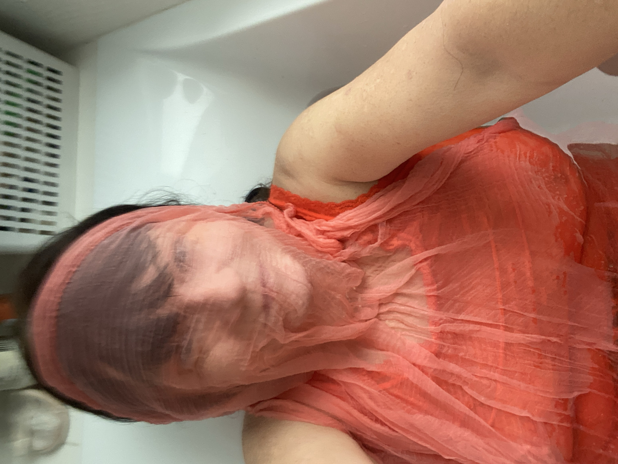 The image shows a person submerged in water, wearing a coral-colored garment. A sheer fabric covers the person's face. The person is in what appears to be a bathtub. The environment suggests an indoor setting. The image does not contain any information that would allow for identification of the location in Novi Petrivtsi, Ukraine.