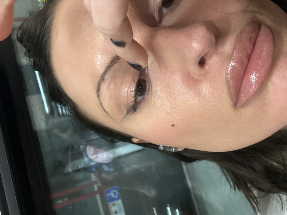 A close-up, angled portrait captures a female individual's face, predominantly from the forehead to the chin. She has dark hair, dark groomed eyebrows with a small red mark visible above the inner corner of the left eyebrow, prominent winged eyeliner, mascara on her eyelashes, brown eyes, and glossy lips. A singular mole is present on her left cheek. Her right hand, featuring fingers with dark nail polish, is positioned near her left eye, specifically touching the lower eyelid. The blurred background, visible through what appears to be a vehicle window, displays reflections and interior elements, including a partially visible blue and white accessibility sticker featuring a wheelchair symbol. The scene is located within a public transport vehicle, consistent with a bus or train, in the city of London, United Kingdom.