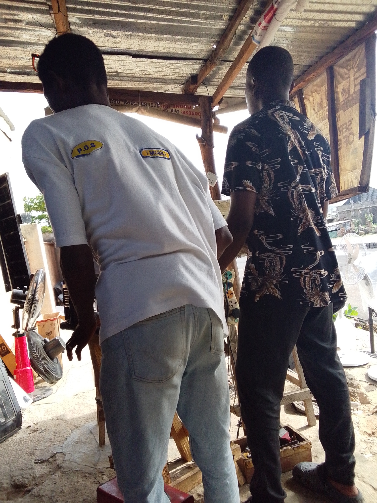 Two men stand under a makeshift structure with a corrugated metal roof and wooden supports. The man on the left wears a white t-shirt with "P.O.S" and "I-MOBILE" labels, blue jeans, and faces away from the camera. The man on the right wears a dark shirt with an ornate pattern, dark pants, and shoes. Several electric fans and other objects are visible. Visible text reads "ADDRESS: Opp." and "PHONE NO: 070-8653898 080" and a number "70" printed on a red object. Based on the OCR text and the background details, this is likely a small business or service point, and the location is the city of Lokoja, Nigeria.