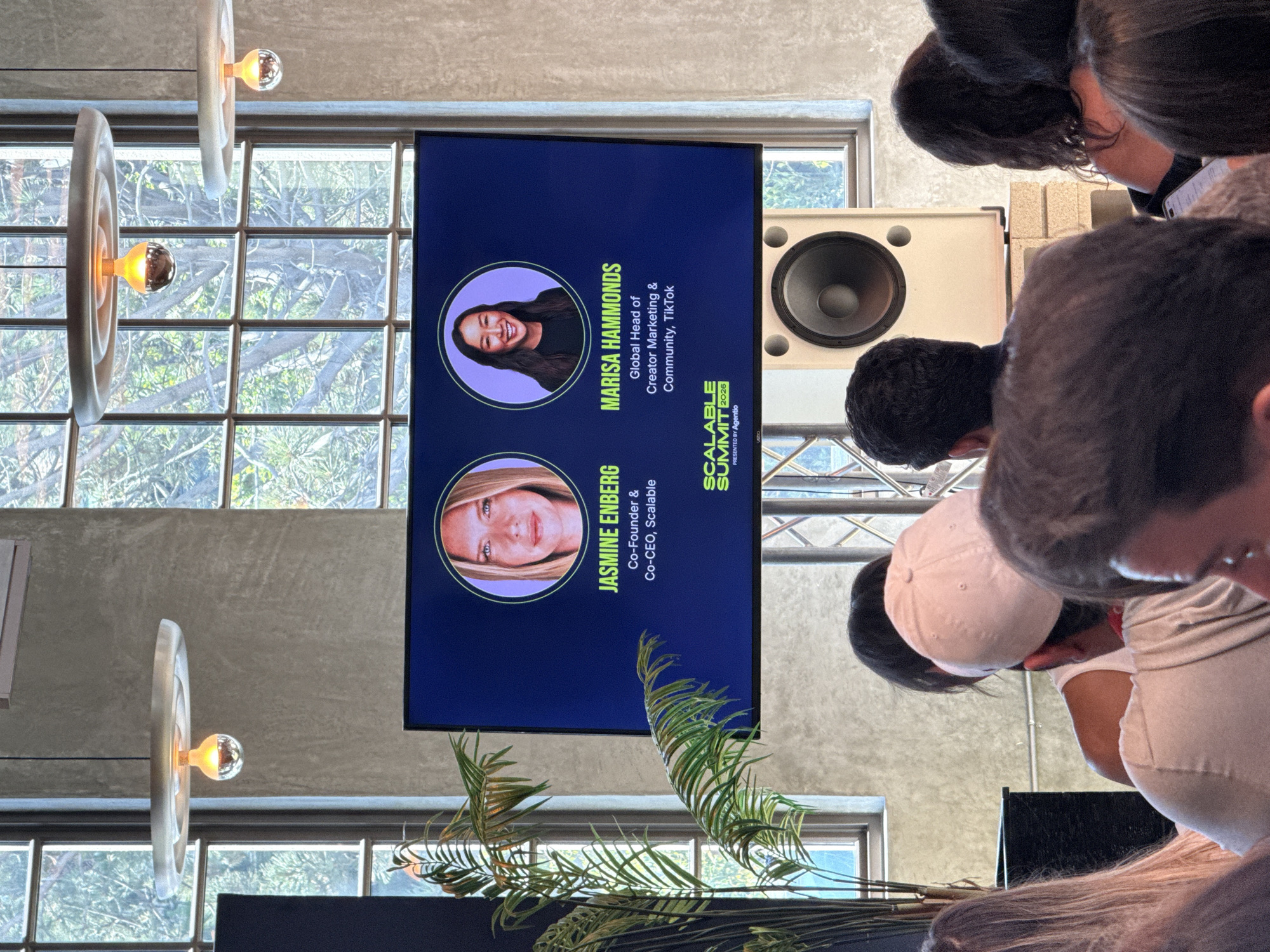 The image depicts a presentation at the "Scalable Summit 2026" conference, featuring Jasmine Enberg (Co-Founder & Co-CEO, Scalable) and Marisa Hammonds (Global Head of Creator Marketing & Community, TikTok). The presentation is displayed on a screen in front of an audience, with a speaker nearby, in a venue that appears to be located in Los Angeles, United States of America. The scene includes a window, lighting fixtures, and decorative plants. The audience members are visible in the foreground, looking at the presentation. There is a large speaker positioned next to the screen.