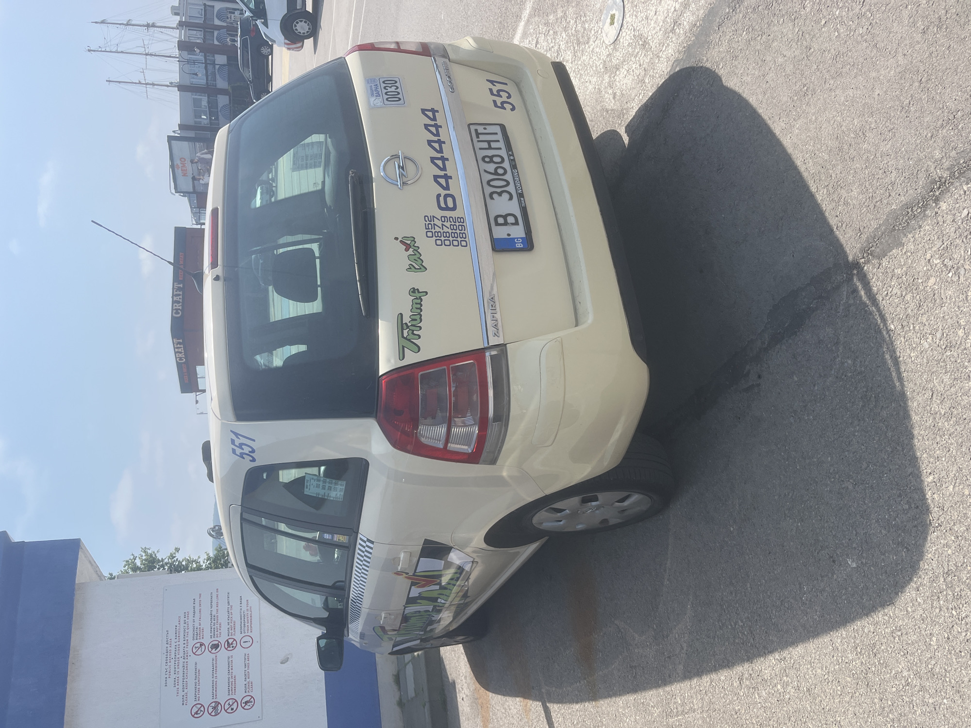An outdoor, high-angle shot captures a light-colored (likely yellow or cream) Opel Zafira taxi parked on asphalt pavement under bright sunlight. The car is angled, showing its rear and part of its left side.
On the back of the taxi, "Triumf taxi" is written in a stylized font, accompanied by a series of phone numbers starting with 052, 087, 088, and 089, all followed by "644444". The taxi also displays the number "551" on its rear window and upper left side. A Bulgarian license plate, "B 3068 HT", is visible, with "TEAM NORDRING BA" written on its frame. A sticker with "VARNA 0030" is also present.
To the left of the taxi, a white and blue wall features a sign with rules and warnings in Bulgarian and English for a "PUBLIC ACCESS AREA". Notable texts include "THIS AREA IS UNDER VIDEO SURVEILLANCE", "DANGER OF FALLING INTO THE WATER", "SWIMMING IS FORBIDDEN", "DO NOT CROSS THE RED LINE ON THE PIER", and "DO NOT PICK UP THE FLOWERS". Several pictograms illustrate these prohibitions.
In the background, buildings are visible, including one with "CRAFT REEF & MEAT" and another with "NEMO" signs. The masts of a tall ship or sailboat can be seen further back, suggesting a port or marina environment, consistent with Varna, Bulgaria. The clear sky and strong shadows indicate a sunny day. The taxi is stationary, with no apparent activity taking place.