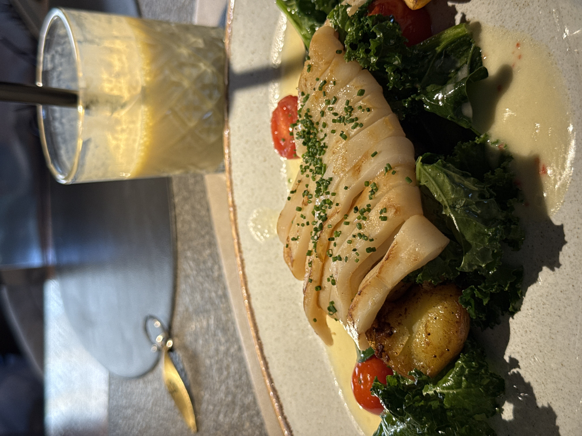 The image shows a plate of grilled calamari, garnished with chives, served on a bed of kale with roasted potatoes and cherry tomatoes. The dish is presented on a grey plate with a creamy sauce. Beside the plate is a glass of a creamy, yellow-colored beverage with a straw. The setting appears to be a restaurant or dining establishment. A silver purse is to the left of the dish. I have no information about the location of the city Krasnoyarsk in Russia.