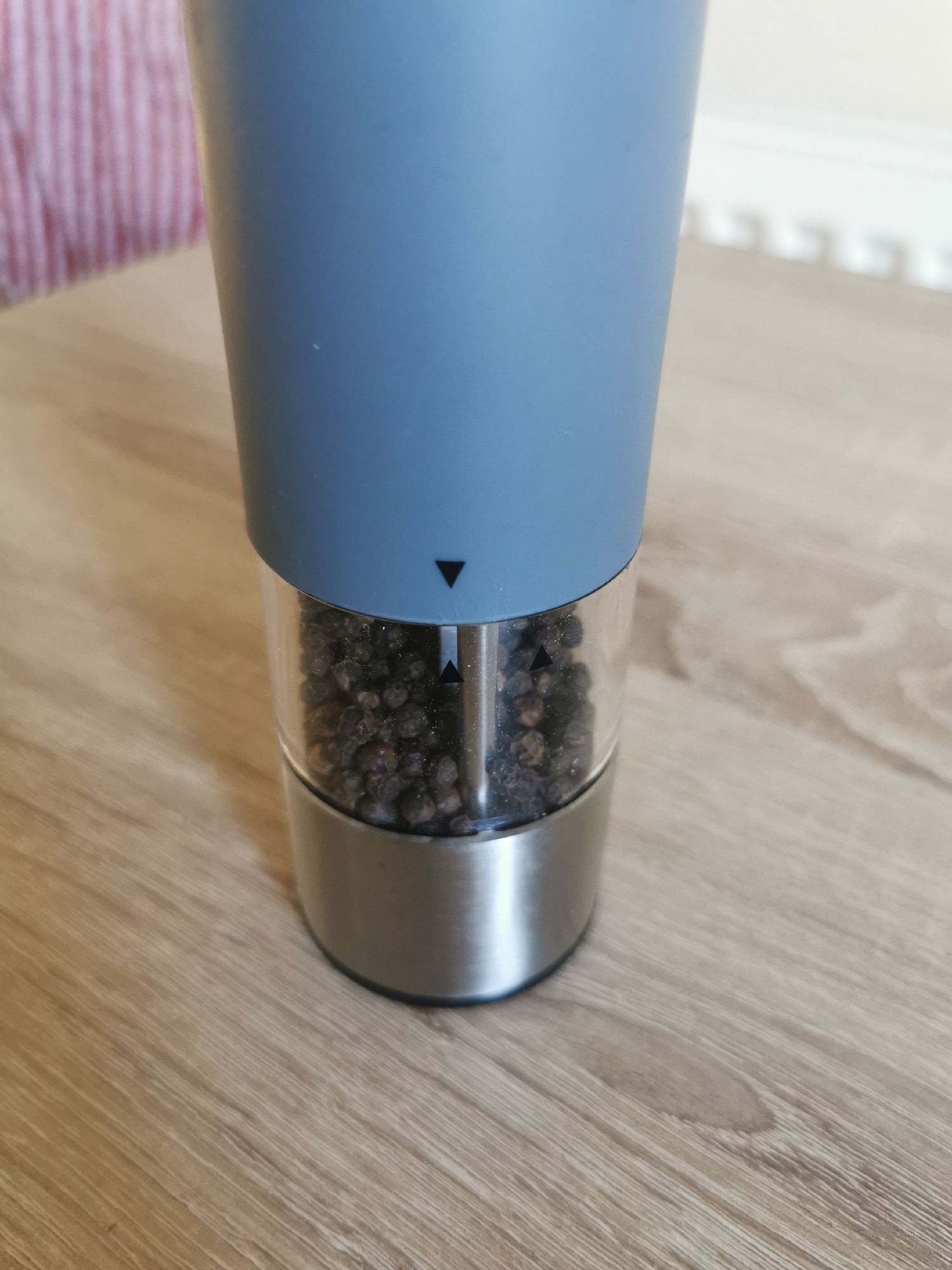 The image shows a close-up view of a pepper grinder containing black peppercorns, resting on a wooden table. The grinder has a blue upper section and a clear glass lower section revealing the contents. There are two small black triangle indicators on the glass part of the grinder, suggesting an adjustment mechanism. The background shows a blurred hint of red and white striped fabric on the left, and a blurred white surface with a radiator-like pattern on the right. The overall scene appears to be indoors, likely a domestic setting. There is no identifiable location in the city of Ripon, United Kingdom.