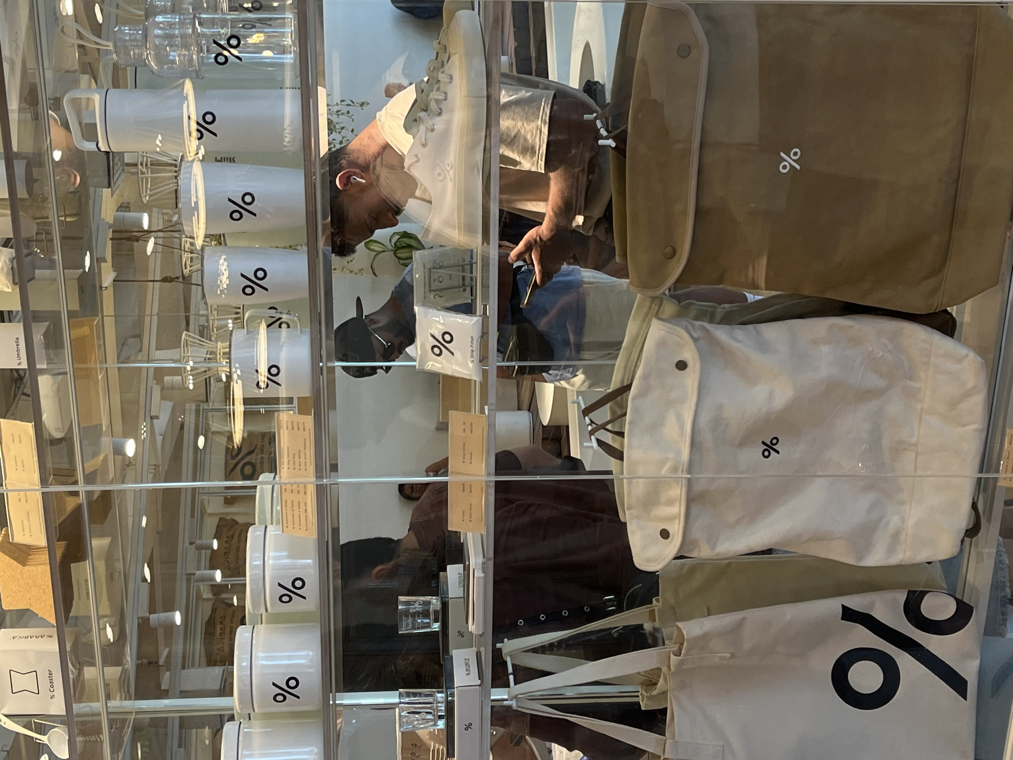 The image shows a retail display case in what appears to be a coffee shop in Kuta, Indonesia. The display case is made of glass and is filled with various products branded with a distinctive "%" logo. On the top shelf, there are white cylindrical containers, some with lids, and clear plastic bottles. One white container prominently displays the "%" logo. Below this, on the middle shelf, are more white containers and some clear glasses, also featuring the "%" logo. A white sneaker with the "%" logo is also visible on this shelf. The bottom shelf showcases several large tote bags and backpacks, all bearing the "%" logo in various sizes and colors, including white and tan. Two men are partially visible behind the display case. One man, wearing a black baseball cap and sunglasses, is looking towards the right. The other man, wearing a light-colored t-shirt and earbuds, is looking down at his phone. The overall environment suggests a modern retail setting, likely a store that sells coffee-related merchandise.