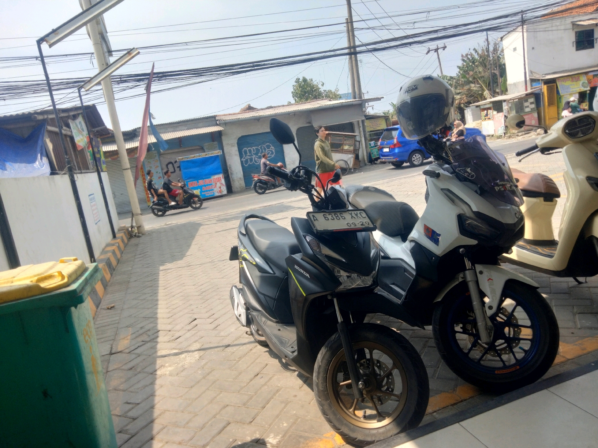 The image shows a street scene in Pisangan Jaya, Indonesia, with several motorcycles parked on a tiled sidewalk. A green trash can with a yellow lid is on the left. In the background, a blue car is parked, and several buildings with corrugated metal roofs line the street. Power lines are visible overhead. Two people are riding a black motorcycle in the middle of the street. Another person in a green jacket and red shorts is walking on the sidewalk. A light-colored scooter is parked in the foreground on the right. A black helmet is resting on the handlebars of a white motorcycle in the center foreground. The license plate of the black motorcycle reads "A 6386 XKC".