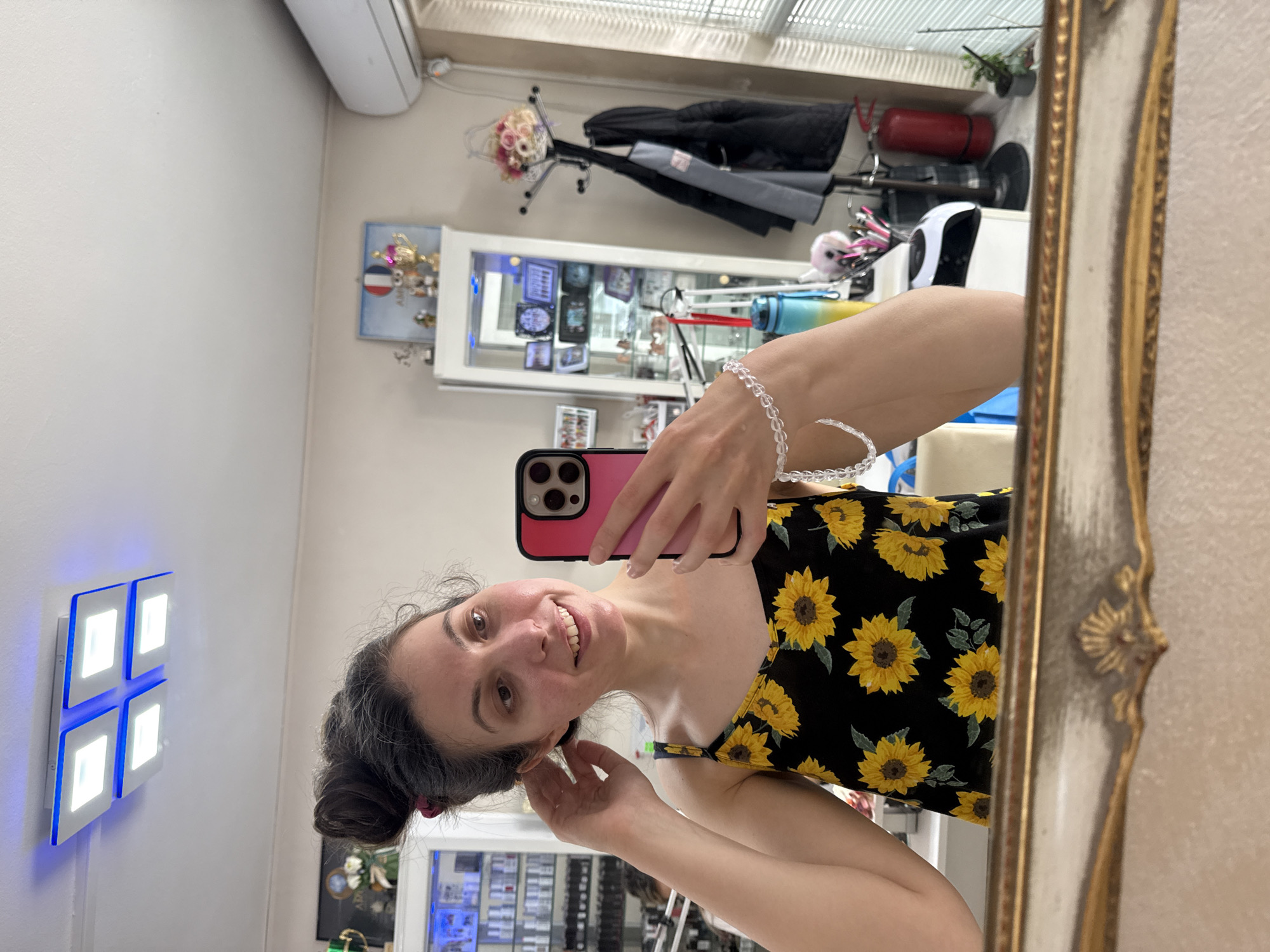 A woman is taking a selfie in a mirror. She is wearing a black dress with a sunflower print and a crystal bracelet. The mirror has an ornate gold frame. In the background, there is a wall with a decorative floral arrangement and a display case filled with various items. The location appears to be a retail establishment. A light fixture is visible on the wall to the left.