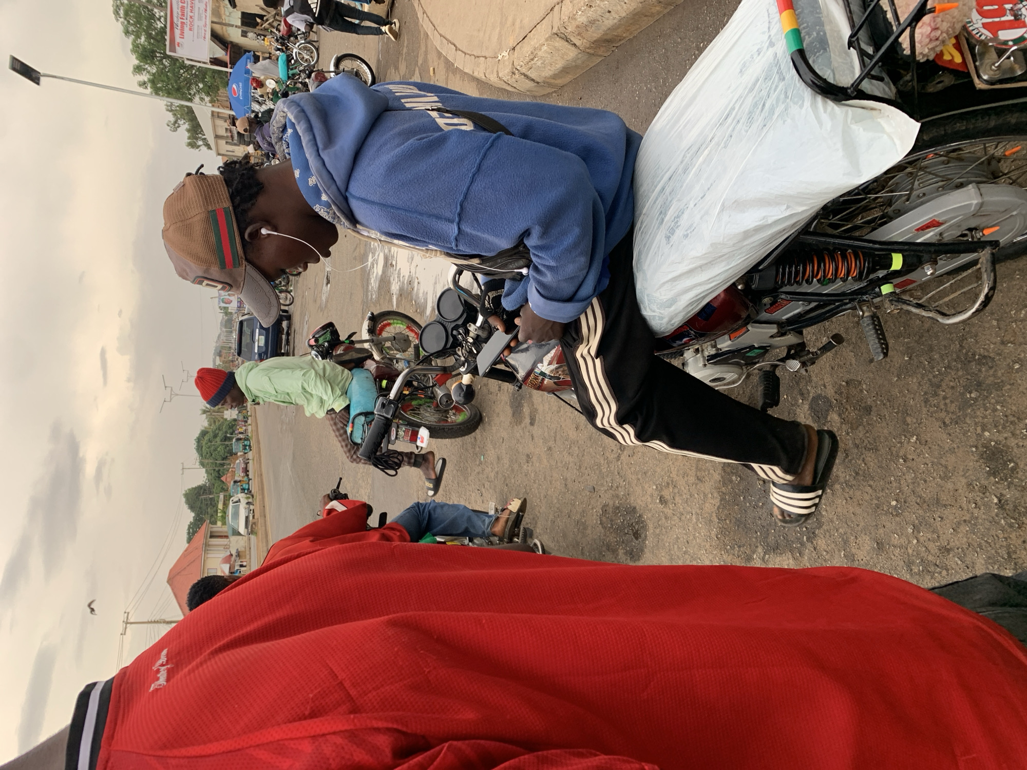 A street scene in New Stadium, Nigeria, shows a row of motorcycles parked along a curb. The perspective is from a low vantage point. A person in a red jacket is in the foreground. A person wearing a blue hoodie, black track pants with white stripes, and a baseball cap sits on one of the motorcycles, looking at a phone in their hands. Another person is on a motorcycle to the left, wearing a light green shirt and a red knit hat. The motorcycles have diverse additions; one has a package wrapped in white plastic attached to the rear and a rack overhead. There are people standing and walking near the motorcycles in the background, which includes shops with signs. The sky is overcast.