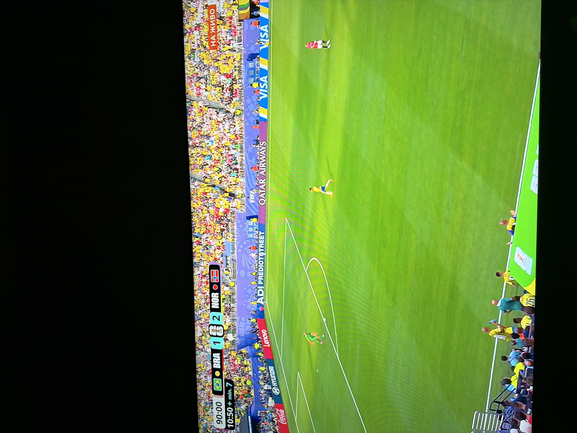 The image shows a soccer match being played on a bright green field under daylight. In the foreground, a player in a green uniform is running with the ball. Further down the field, another player in a red uniform is standing. The background is filled with spectators in a stadium, many wearing yellow clothing, cheering. A scoreboard at the top of the frame displays "90:00 BRA 1 2 NOR" and "10:50 + min. 7". Advertising banners for brands like Coca-Cola, Hyundai, Lenovo, Qatar Airways, and Visa are visible around the perimeter of the field. The overall scene depicts an active sports event. There is no indication of the location being Ognianovo, Bulgaria, within the image itself.