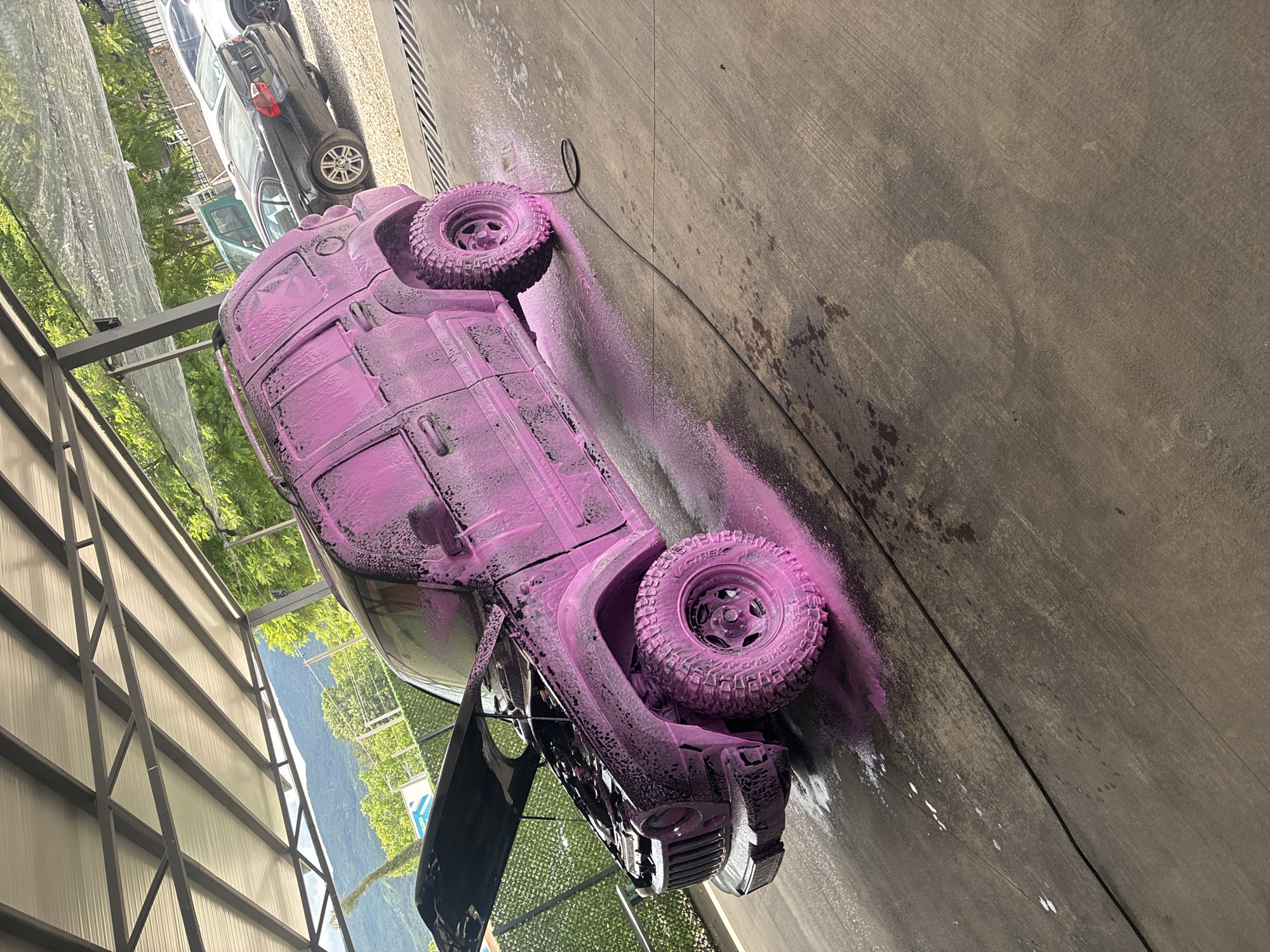 The image shows a black off-road vehicle covered in pink foam, likely during a car wash process. The vehicle is parked on a concrete surface. The car is oriented upside down in the image. Other cars are parked in the background. The location appears to be outdoors, possibly in a car wash facility or similar commercial area. A cable lays on the concrete.