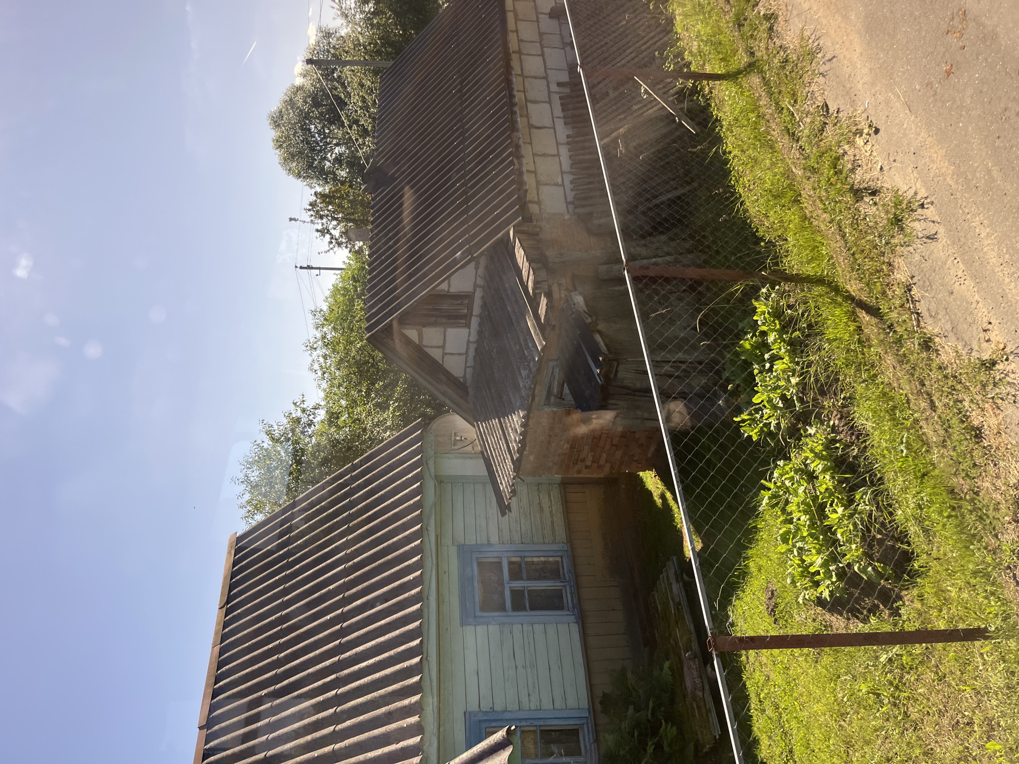 This is a medium-angle, sunlit exterior shot of a rural property in Pastavy, Belarus. The foreground features a chain-link fence with rusted metal posts, enclosing a patch of green grass and a small garden bed with leafy plants. Beyond the fence, two buildings are visible. The building on the left is a light-blue, wooden house with a dark, corrugated roof and a light-yellow foundation. It has two windows with blue trim. Adjacent to it and slightly behind is a smaller, brick structure with a corrugated roof, appearing to be an extension or outbuilding. The background is filled with green trees and a clear blue sky with a few wispy clouds. The scene suggests a residential property in a rural or suburban setting.