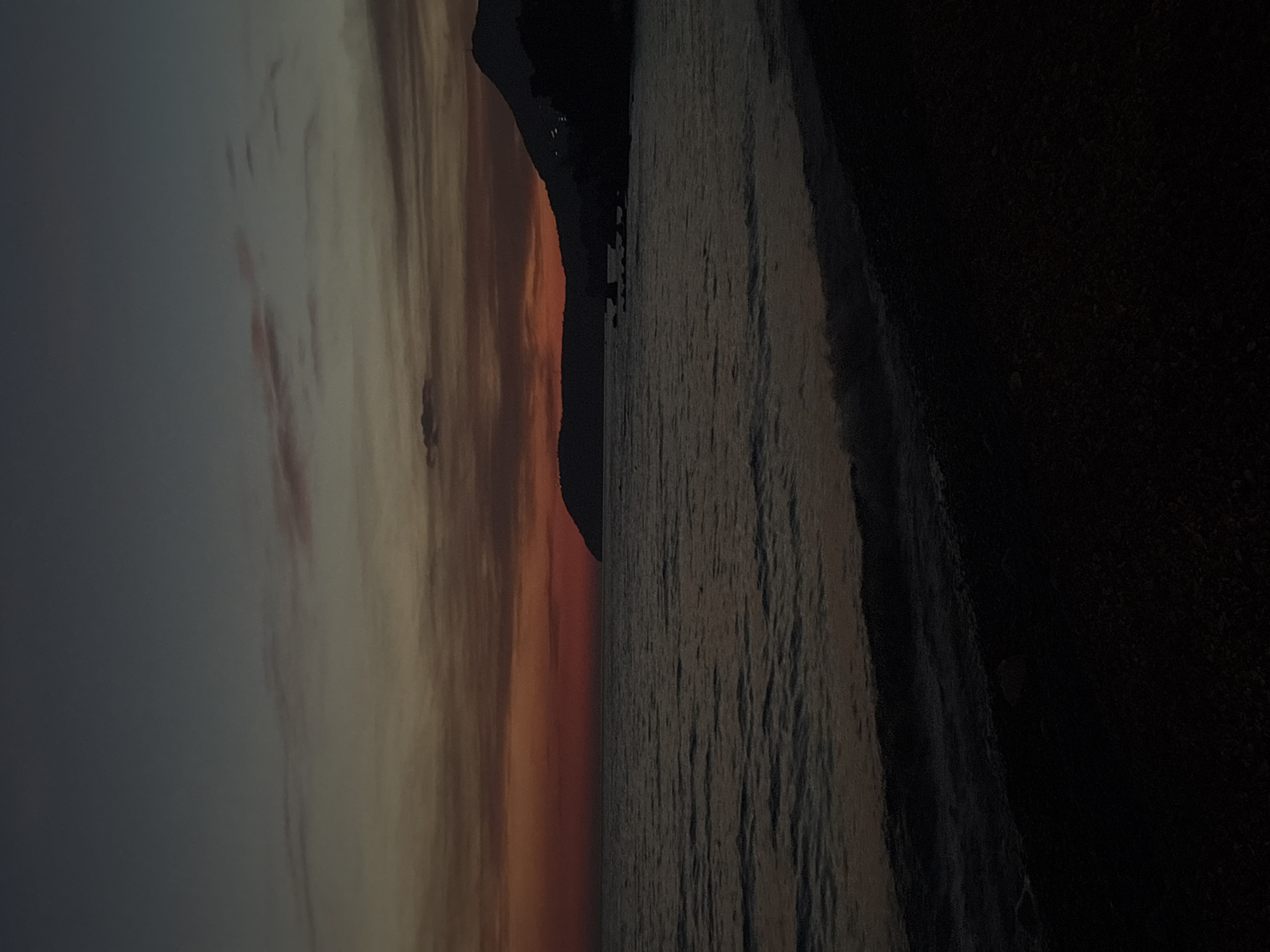 The image shows a sunset over the Adriatic Sea in Bar, Montenegro. The sky transitions from light gray to orange near the horizon, with clouds visible. A dark, silhouetted land mass is visible between the sky and the water. The sea is a dark gray color. A dark, unidentifiable area is in the foreground on the right.
