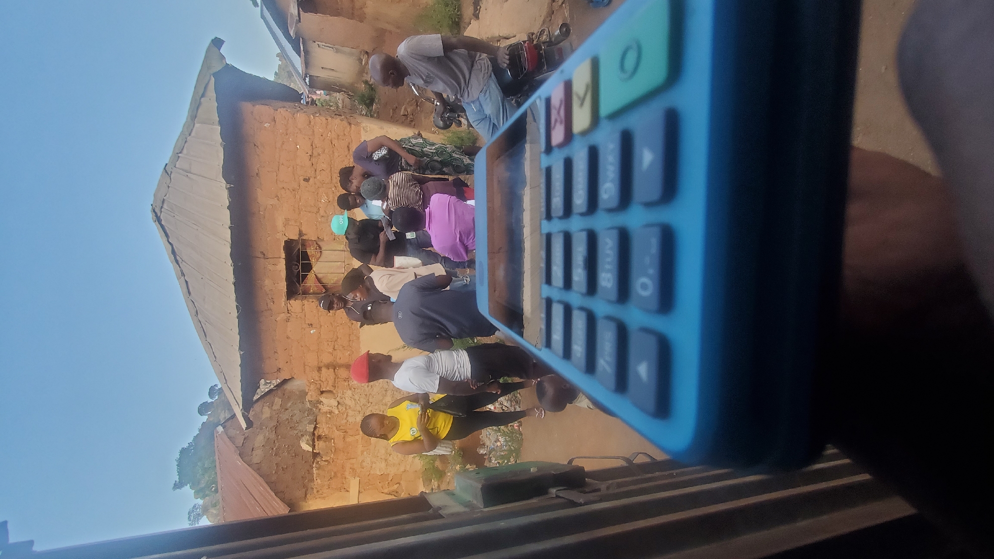 The image shows a group of approximately 10-15 people gathered outside a building in Vom, Nigeria. The building appears to be constructed of mud bricks and has a corrugated metal roof. A motorcycle is parked near the group. In the foreground, a blue mobile point-of-sale (POS) device is prominently displayed, obscuring part of the scene. The lighting suggests it is daytime. Several individuals are visible, their actions and interactions are obscured by the POS device.