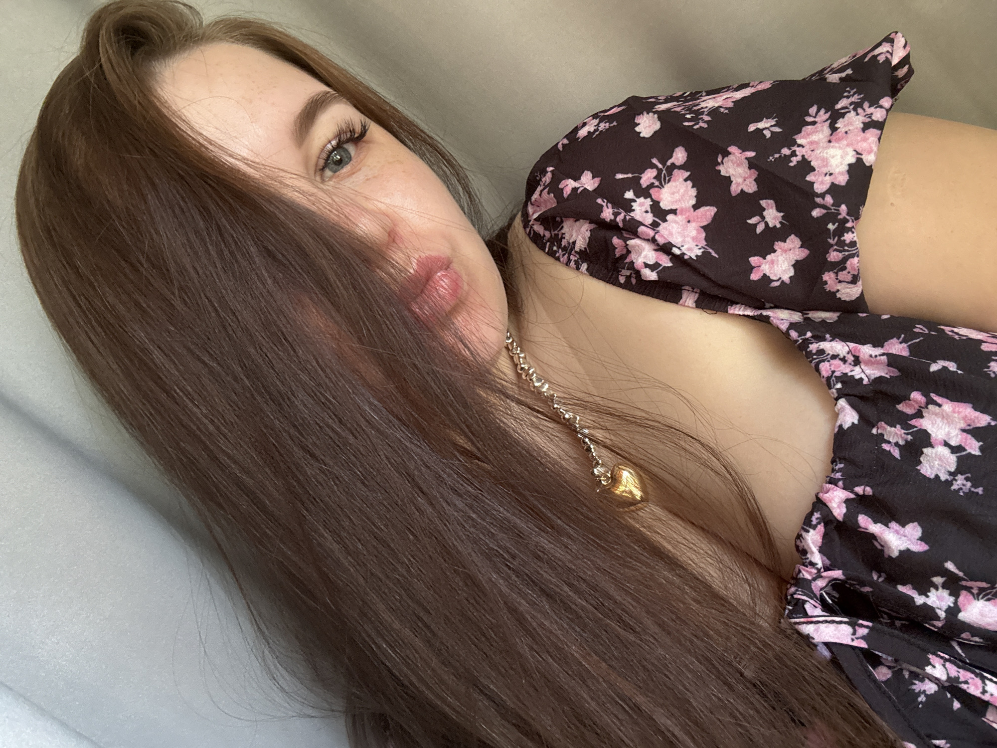 The media file is a close-up, self-portrait of a young woman. She has long, dark brown hair that partially obscures her face, covering her left eye and part of her forehead. Her right eye, which is visible, is light blue with long, dark eyelashes and appears to be looking towards the camera. She has fair skin with light freckles visible on her cheeks. Her lips are slightly parted, and she is wearing a dark, floral-patterned dress with pink flowers. A delicate silver necklace with a gold heart-shaped pendant is visible against her neck and chest. The background is a neutral, light-colored surface, possibly a sofa or bed. The lighting suggests an indoor setting, likely during the daytime. The overall mood is relaxed and intimate. There is no visible text in the image. The location is identified as Chaikovsky, Russia.