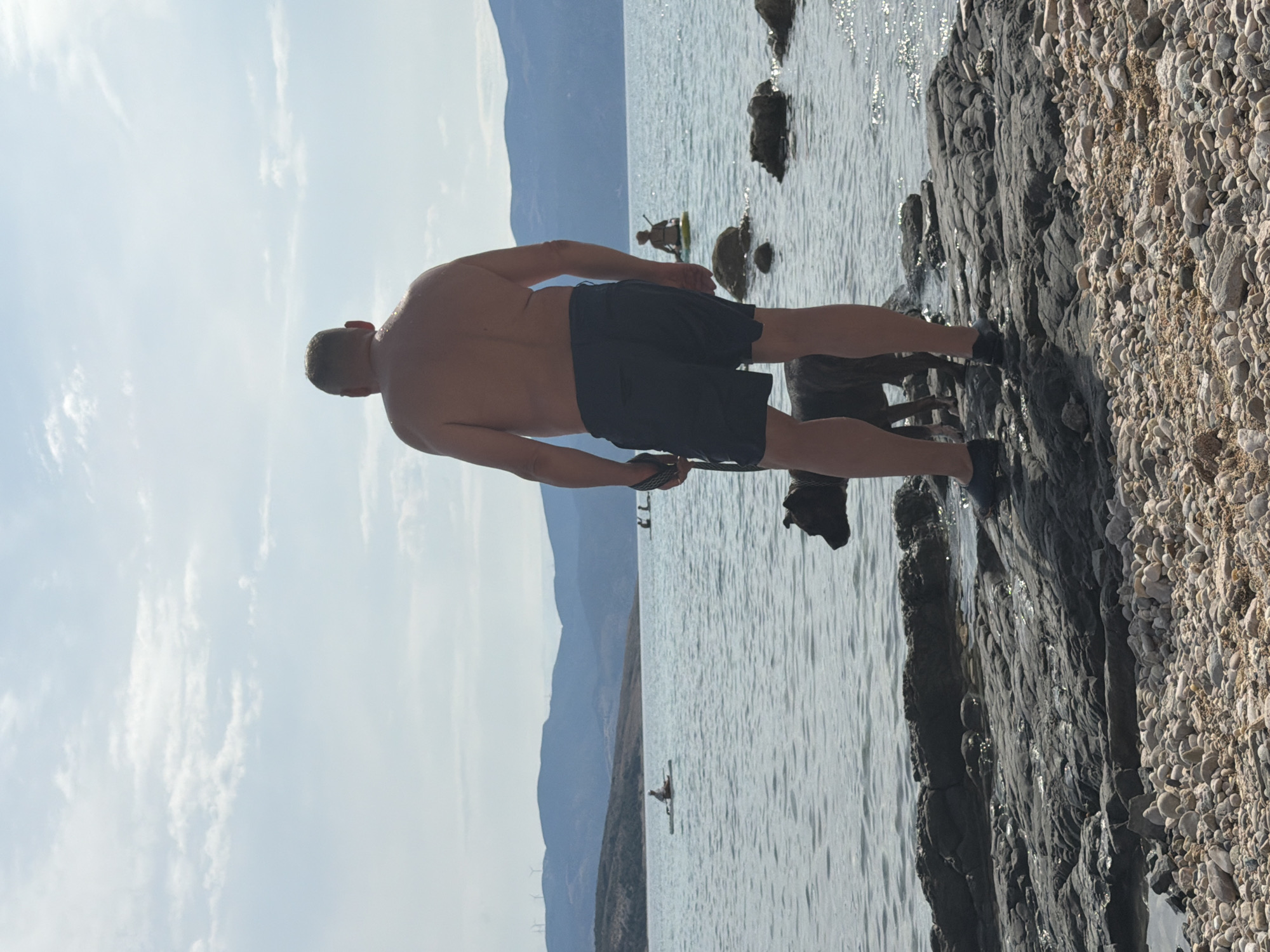 In Općina Baška, Croatia, a shirtless man stands on a rocky shore, facing away from the camera, looking out at a calm body of water. He is wearing dark shorts and dark shoes. Beside him, a dark-colored dog on a leash also faces the water. In the background, several people are seen on paddleboards in the water, and a hazy, mountainous landscape borders the horizon under a partially cloudy sky. The foreground is composed of pebbles and larger rocks.