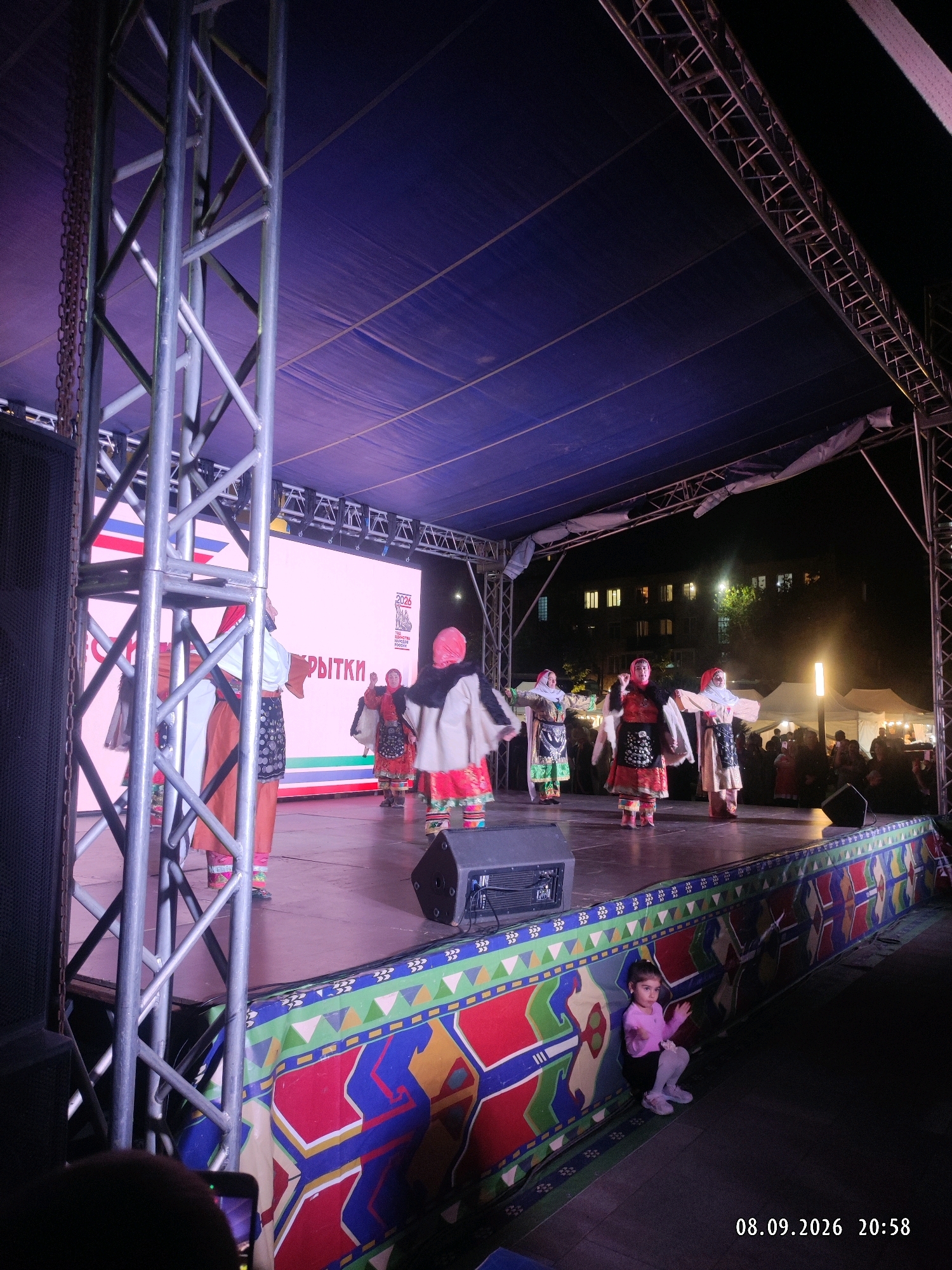On a stage set up outdoors, several people are performing a dance. They are dressed in traditional, colorful clothing, some with headscarves. The stage is covered by a large blue canopy, supported by metal scaffolding. A large screen is visible behind the performers, displaying text in a language that appears to be Russian, and a logo with the year "2026." In front of the stage, a young girl in a pink shirt and white leggings is sitting on the ground, watching the performance. The surrounding area suggests an evening event, with artificial lighting illuminating the stage and some buildings visible in the background. The overall scene is festive and cultural.