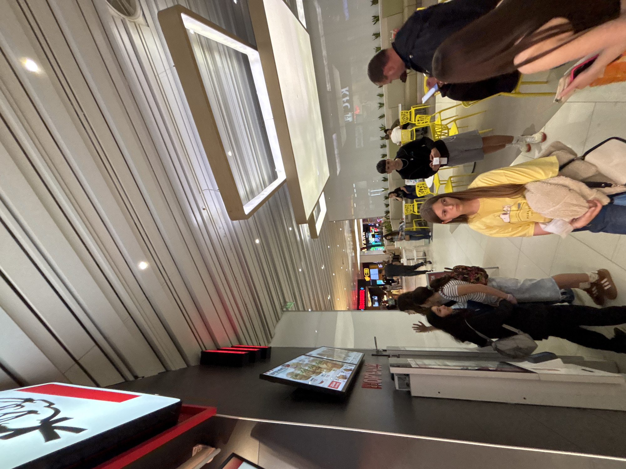 The image shows a busy scene inside a shopping mall, possibly in Plovdiv, Bulgaria (if location is required). The mall has a high ceiling with parallel linear lighting features. In the foreground, there's a KFC sign. Further into the scene, there is a display case with fast food menu. Several people are visible: Some are seated at yellow tables and chairs. In the middle ground, a young woman with long brown hair is wearing a yellow shirt. Other people include couples, families, and individuals walking around the mall. The environment is generally brightly lit and appears to be a busy day at the mall.