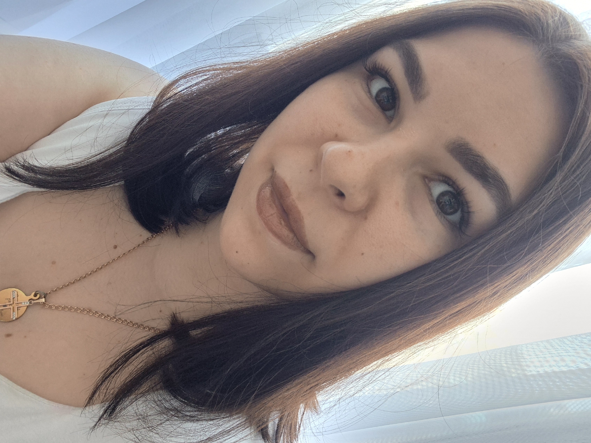 A close-up, slightly angled selfie depicts a young woman with brown hair and eyes. She wears light makeup, including eyebrow shaping and lip color. A gold chain necklace with a pendant is visible around her neck. The background appears to be a window with white sheer curtains. There is no information to suggest this image was taken in Tobolsk, Russia.