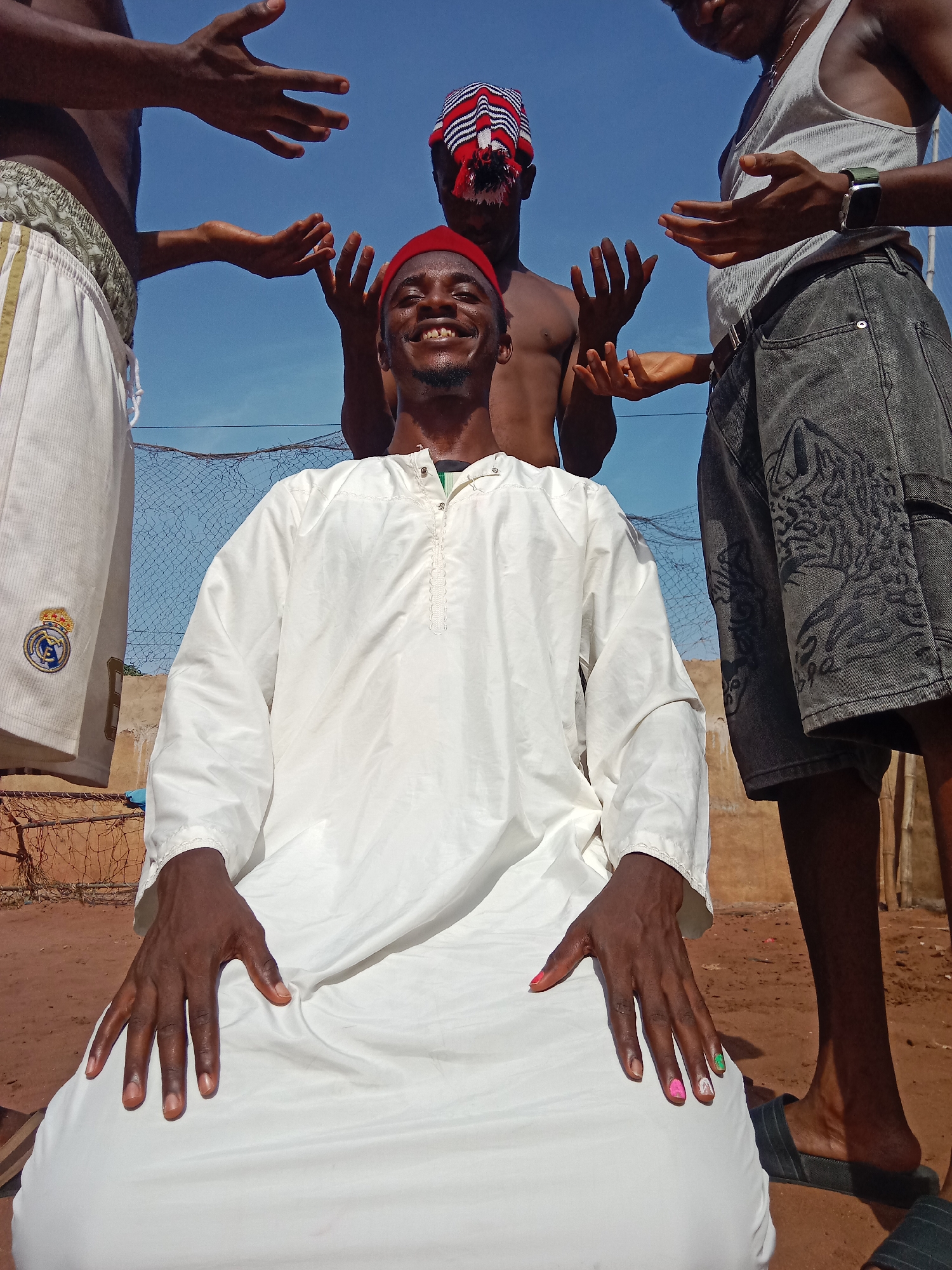 A medium eye-level shot shows a dark-skinned man kneeling with three other men standing behind him. The kneeling man is wearing a long white robe with a decorative stitch pattern along the upper chest area and a red cap. He is smiling broadly. His fingernails are painted in various colors. The man directly behind him is bare-chested, and wearing a striped headscarf. The two men flanking him are both partially cropped out of the image. The man to the left is wearing white shorts with a Real Madrid soccer logo. The man on the right is wearing a grey tank top and patterned denim shorts. All three men behind the kneeling man have their hands raised, palms facing upward. A blue sky is visible above the men. A dilapidated fence constructed of netting and wooden sticks is visible behind the group. The ground is red dirt. The location is the city of Unknown, Unknown.