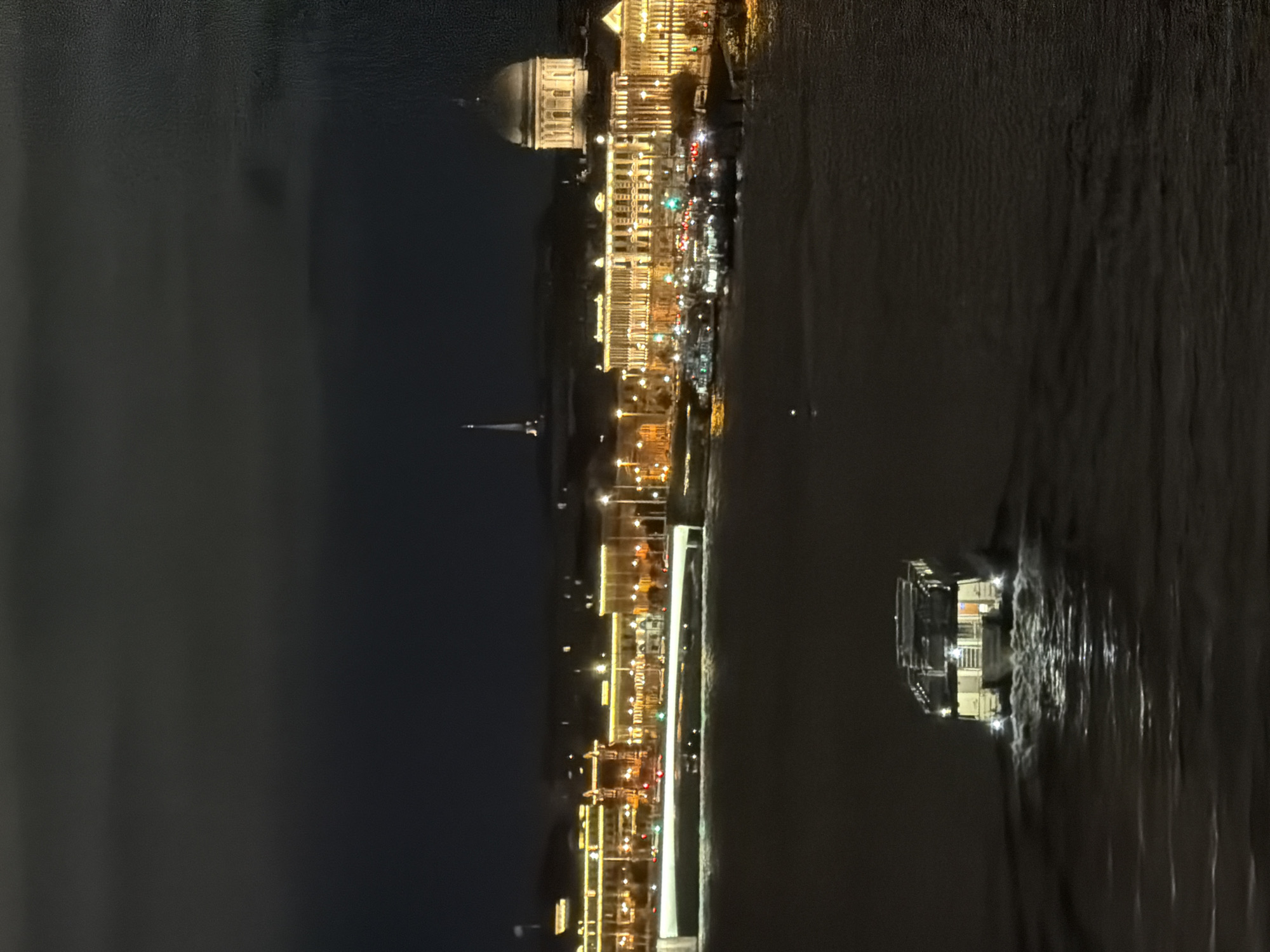 The image depicts a nighttime view of the Saint Petersburg skyline, likely from a boat on the Neva River. In the foreground, a white boat with illuminated windows is moving away from the viewer, leaving a wake in the dark water. Across the river, a brightly lit cityscape stretches along the embankment. Several multi-story buildings with ornate facades are visible, adorned with numerous yellow lights. A prominent bridge with illuminated railings spans the river in the middle ground. In the background, a tall, slender spire rises into the dark sky, and a large building with a dome is visible on the right. The sky is dark and cloudy, with the city lights reflecting on the water's surface.