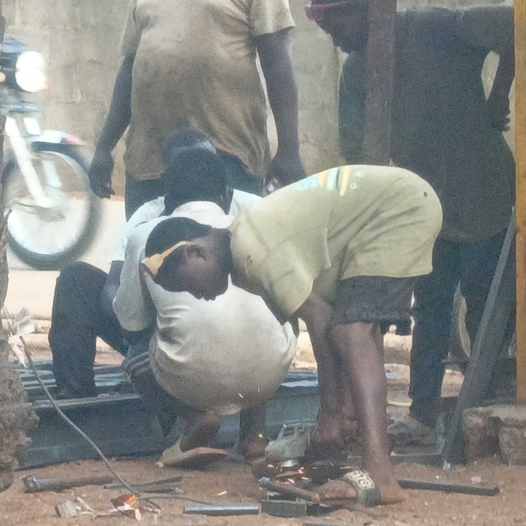 This full-shot image captures several men engaged in metalworking outdoors in what appears to be an informal workshop in Akwanga, Nigeria.
In the foreground, a man wearing a light-colored t-shirt and dark shorts is bent over, operating a power tool, likely an angle grinder or welding equipment, as indicated by the visible sparks emanating from the metal pieces on the ground. He is wearing sandals, with the word "BIG" faintly visible on the strap of his left sandal. Another man in a white shirt is crouching or sitting directly behind him, observing or possibly assisting.
Further back on the left, a man in a light t-shirt stands, with only his torso and arm visible. On the far right, another person in dark clothing is partially seen, possibly standing. The men appear to be working on metal bars or frames scattered on the dusty ground, with a wire or cable running across the work area.
In the blurred background on the left, the front wheel and partial body of a motorcycle or scooter are visible. The background also features a light-colored brick or concrete wall. The activity is taking place during the daytime. A notable detail is the apparent lack of personal protective equipment (PPE) such as safety glasses or a welding mask for the man operating the spark-producing tool. The overall scene depicts informal industrial or craft work.