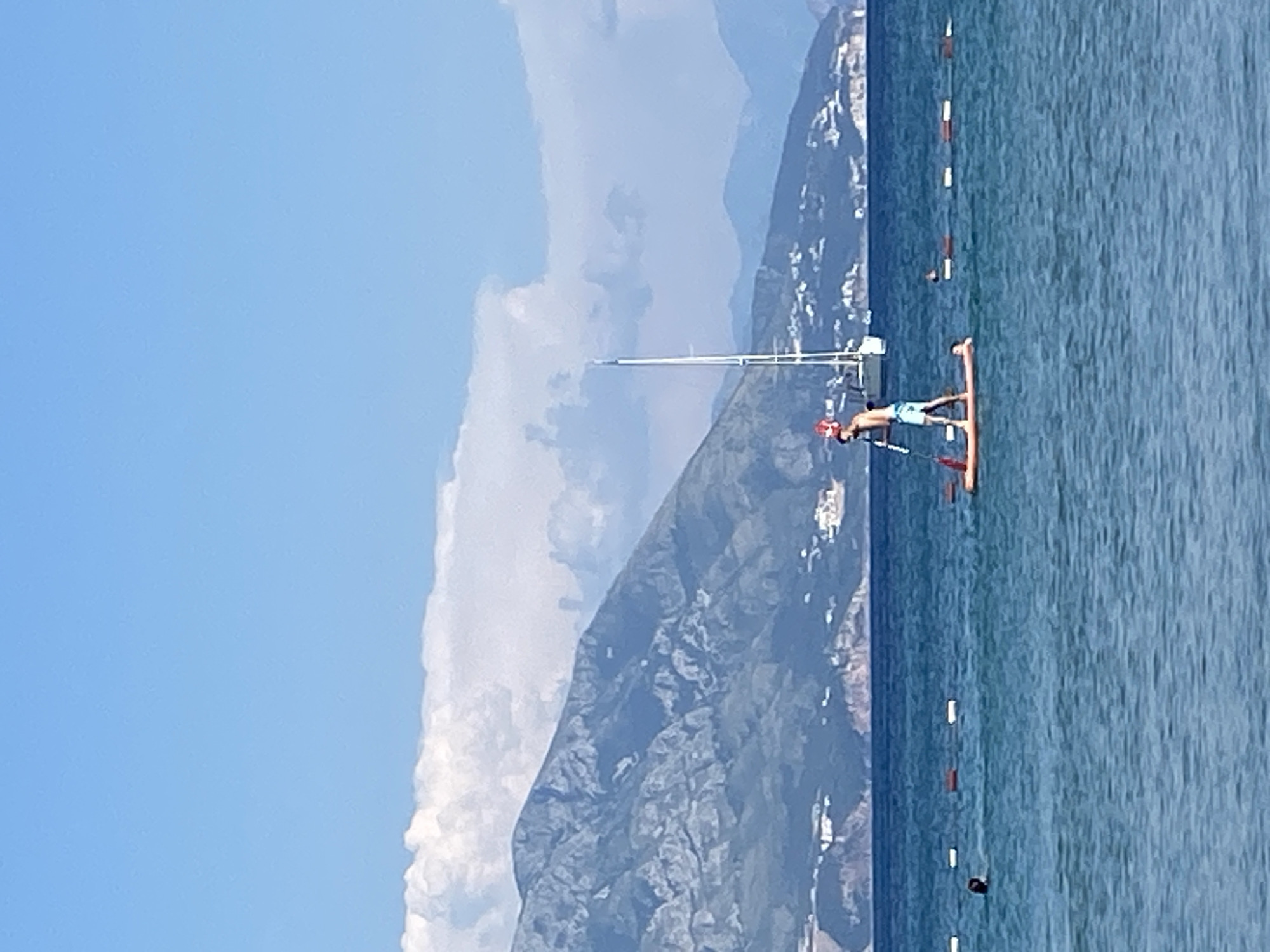 The image shows a person stand-up paddleboarding on a calm sea under a clear blue sky with some clouds. In the background, there are mountains with some buildings on their slopes, and a sailboat anchored offshore. The water is a deep blue, and there are buoys marking a swimming area. The paddleboarder is wearing blue shorts and appears to be holding a paddle. The person is positioned in the center-left of the frame. The scene is likely on the coast of Budva, Montenegro, known for its scenic mountain backdrop and coastal activities.