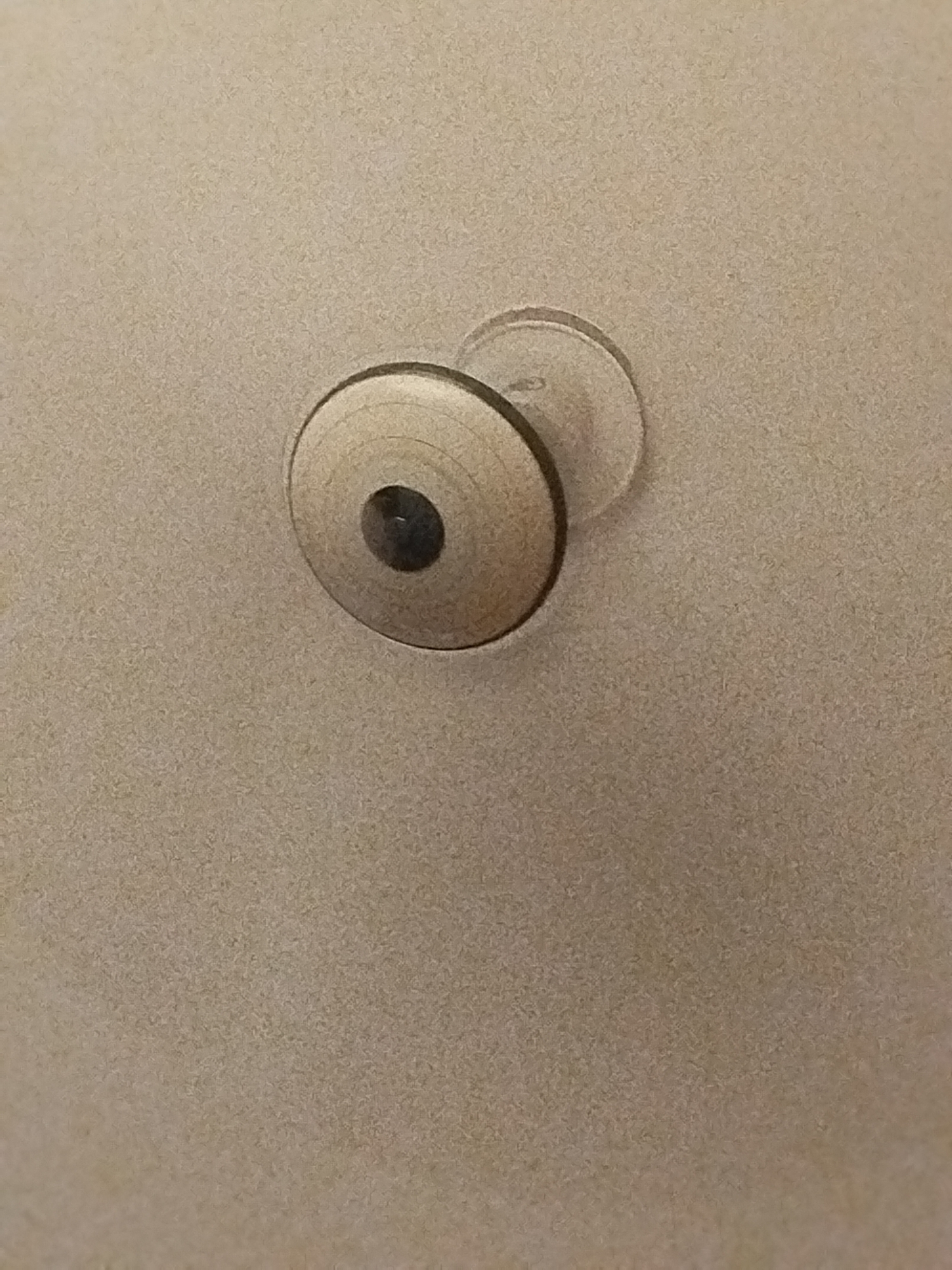 The image displays a close-up view of a silver, circular object attached to a textured, beige surface. The object appears to be a knob or a decorative element, with concentric rings etched into its surface. At the center of the knob, there is a dark, circular indentation, possibly a screw head or a keyhole. A clear, circular backing is visible behind the knob, suggesting it is affixed to the surface. The background is uniformly textured and appears to be a wall or a ceiling. There is no visible indication of location in Lassan, Pakistan.