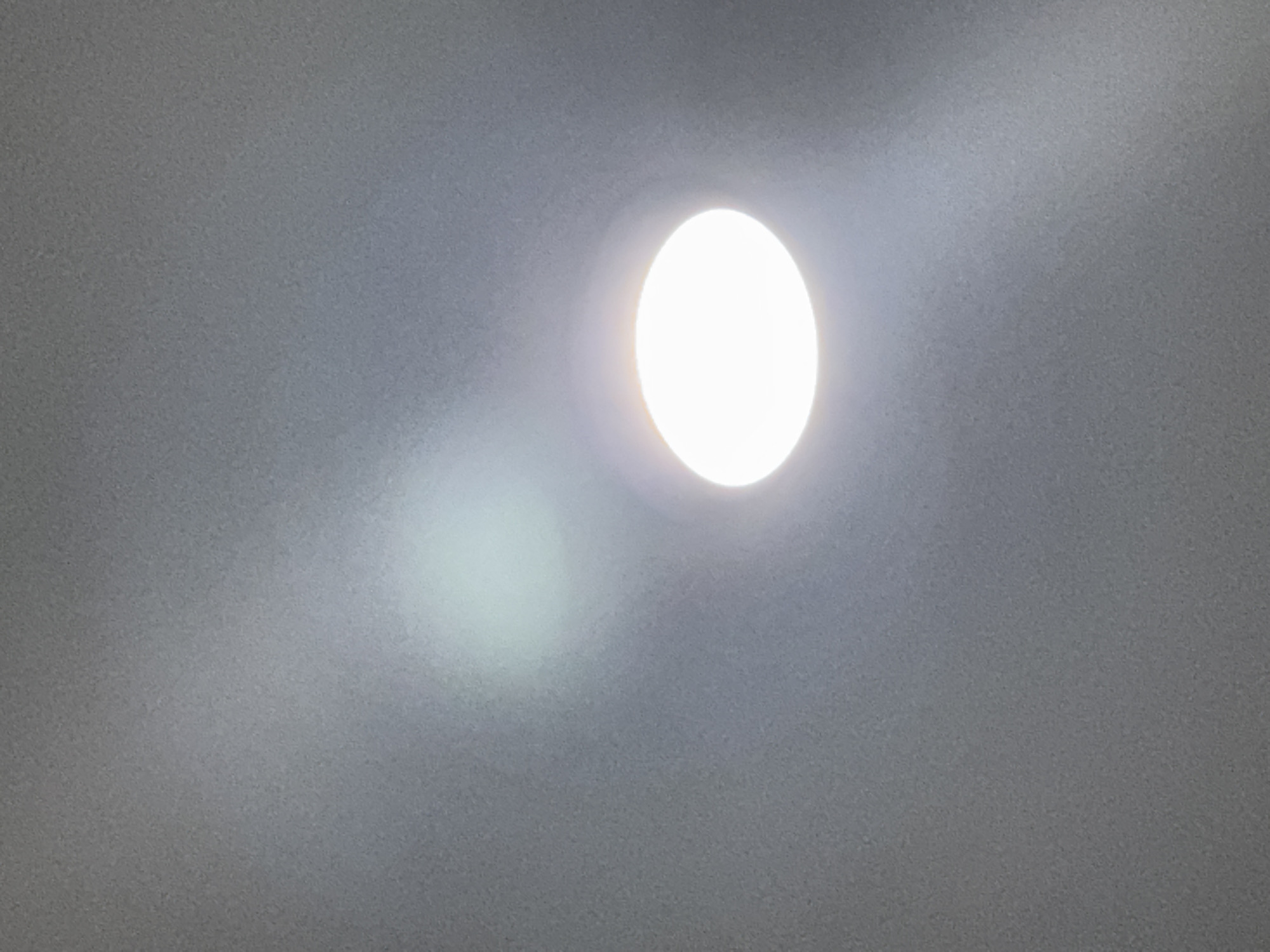 An overhead, close-up shot captures a bright, circular light fixture against a textured, gray ceiling. The light is intensely white and appears to be a recessed LED downlight, creating a circular halo of illumination. To the left of the light, a softer, less defined luminous patch suggests a reflection or another light source. The ceiling surface is uniformly gray with a subtle, granular texture. The image appears to be taken indoors, possibly in a residential or commercial space. There are no people, animals, or other objects visible. The scene is static and well-lit.