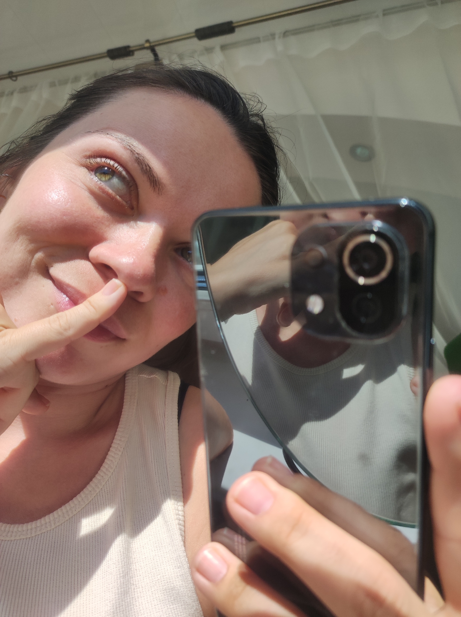 A young woman is taking a selfie with her smartphone. She is wearing a white tank top. She is holding the phone with her right hand and pointing to her nose with her left index finger. The phone's reflective surface shows a blurred reflection of her upper body. The setting appears to be indoors, likely a bright room with white curtains. There is no evidence or information indicating that the photo was taken in the city of Chaikovsky, Russia.
